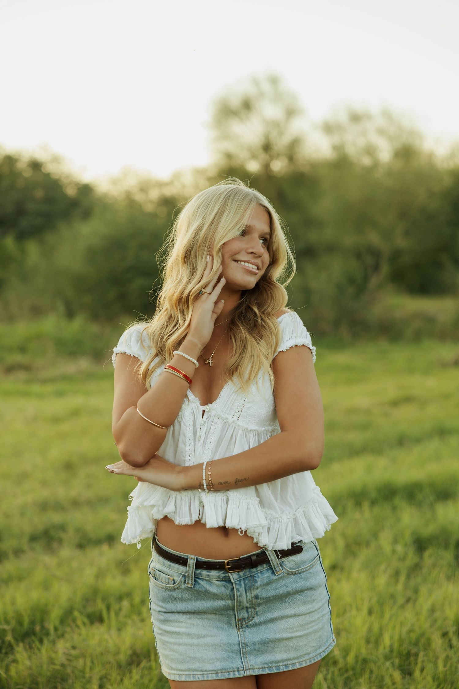 A person in a white crop top and denim skirt poses playfully in a sunny green field. Senior photos