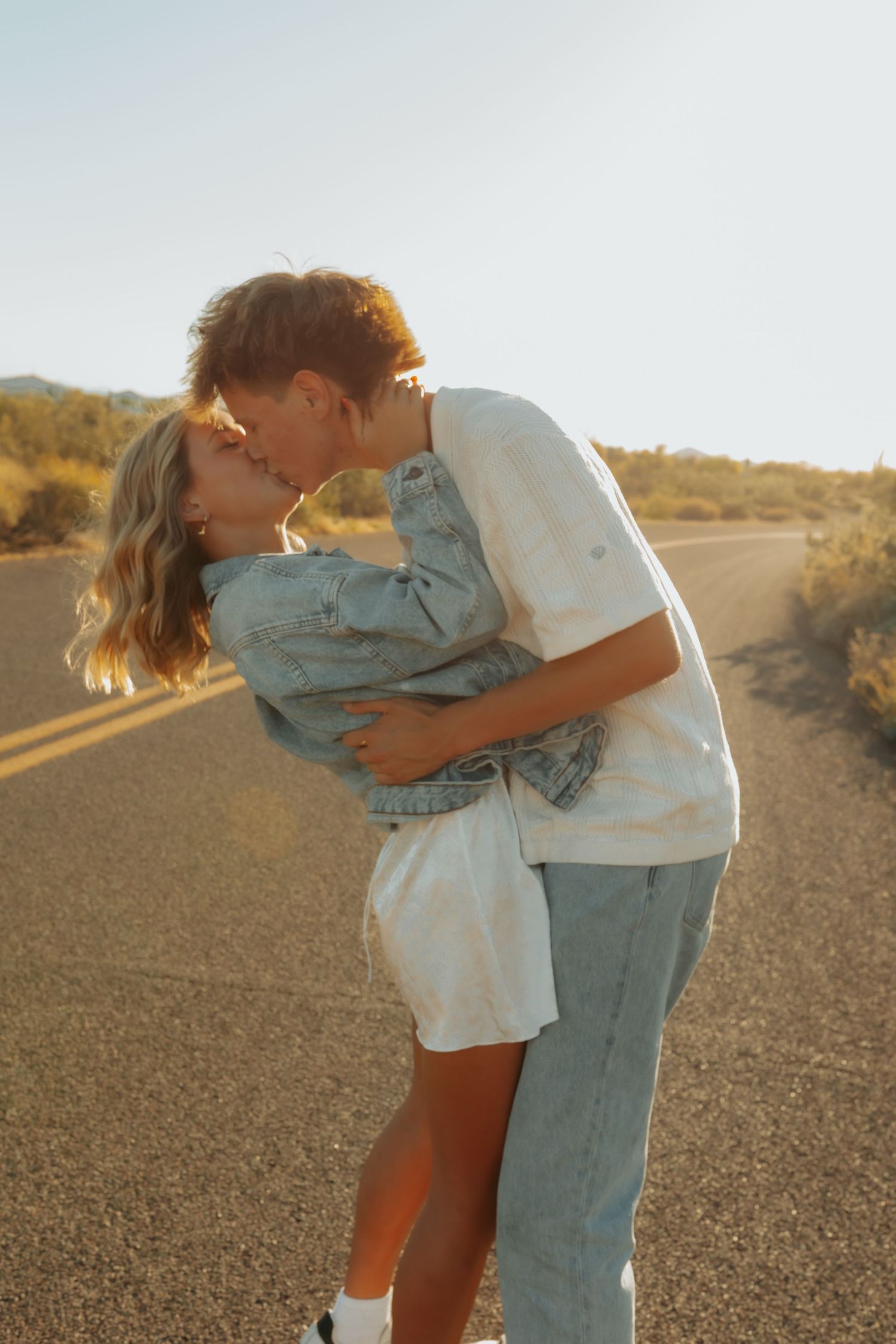 A romantic couple embraces and kisses on a desert road at golden hour with warm sunlight illuminating the scene.