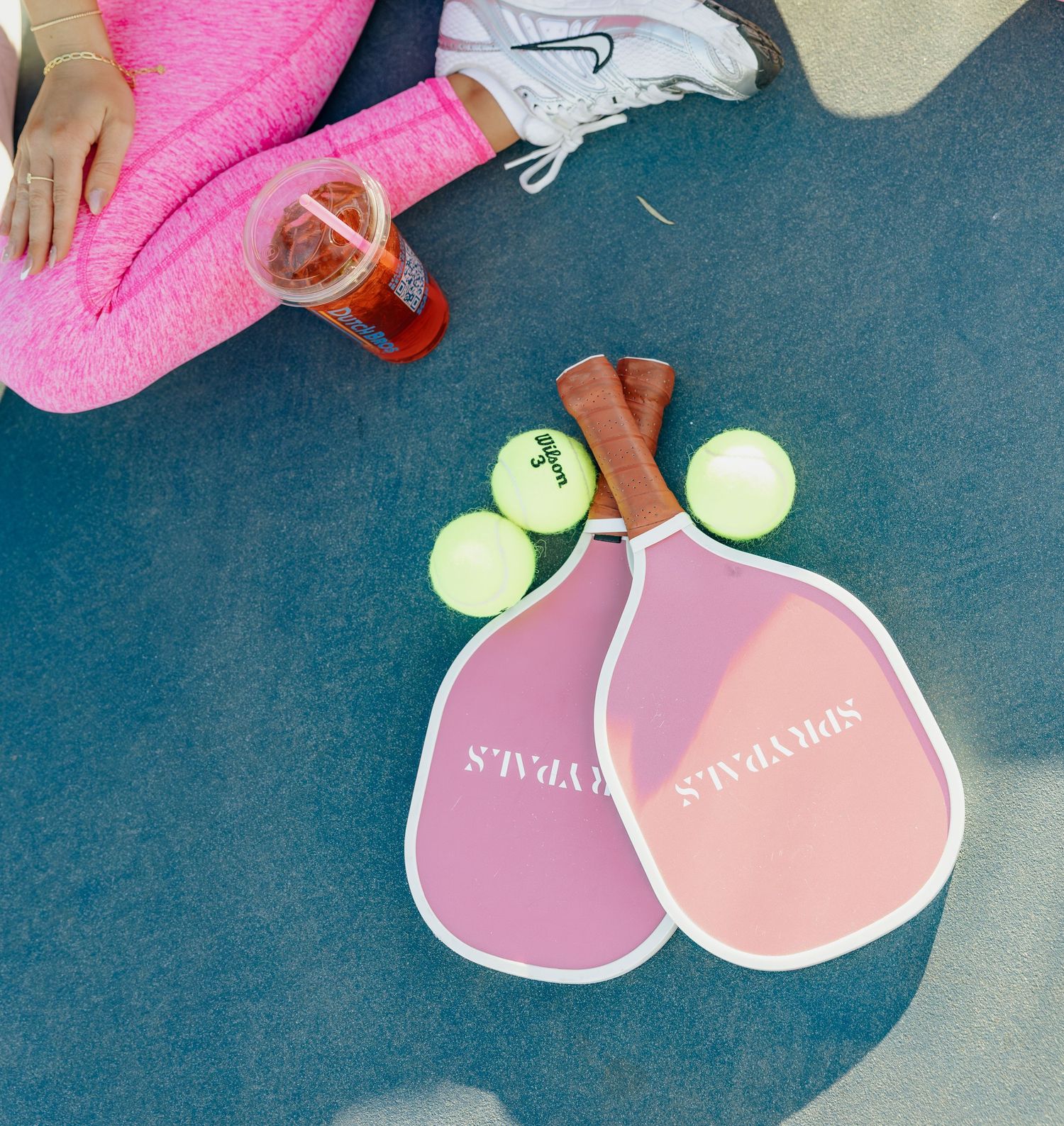 Pink pickleball paddles and tennis balls arranged on blue court surface with a red drink visible in frame.