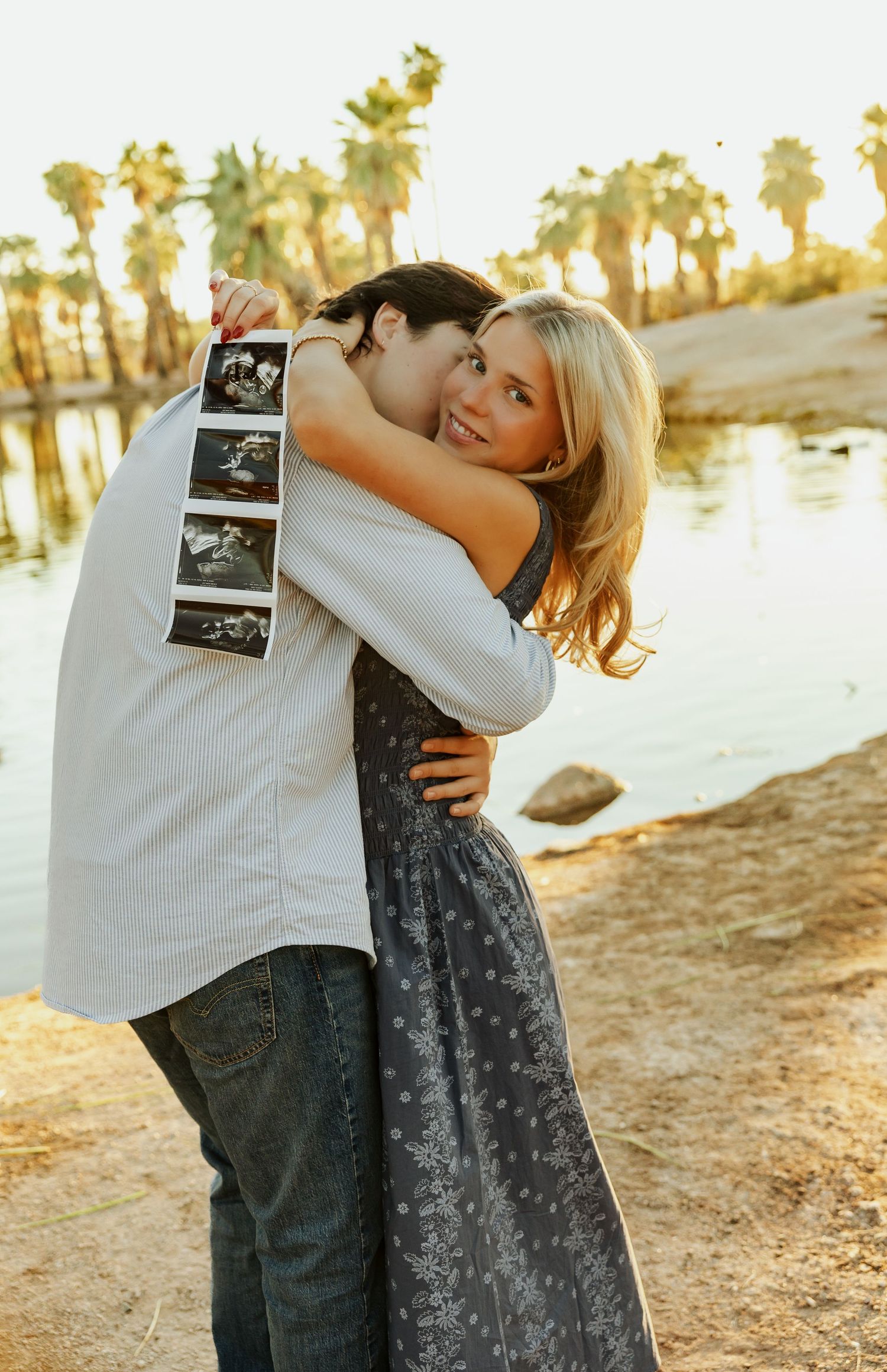 Couple embraces outdoors while holding ultrasound photos during sunset at a lakeside location.