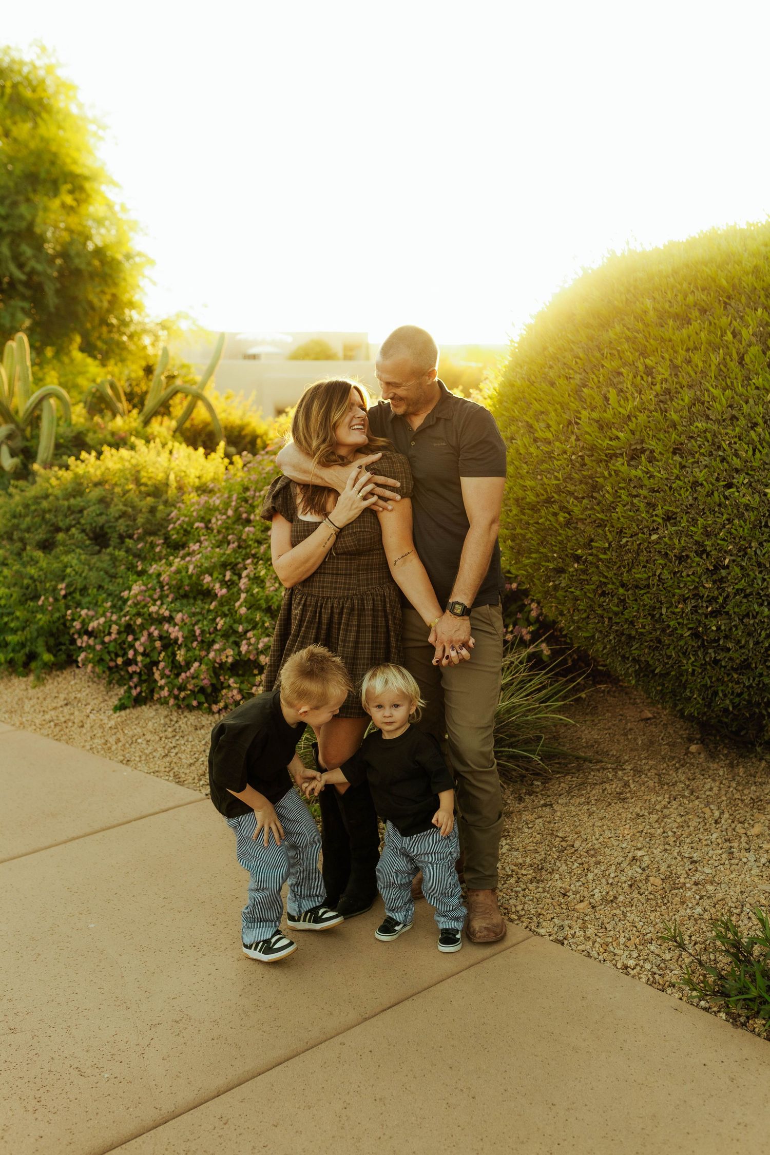 Family of four embraces during golden hour sunset in desert garden with lush greenery and pathway.