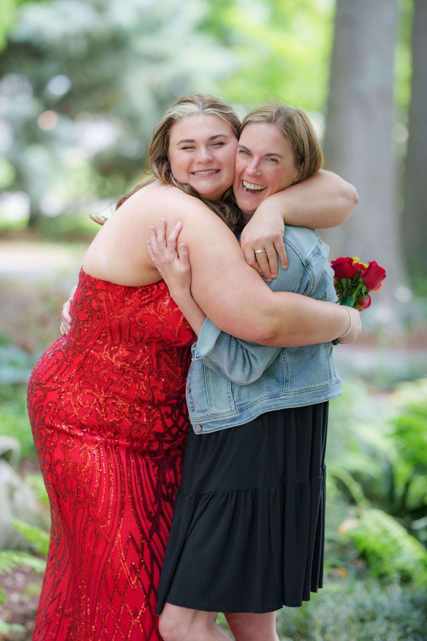 Two women hugging outdoors, one in a red sequin dress holding roses, smiling joyfully in a garden setting.