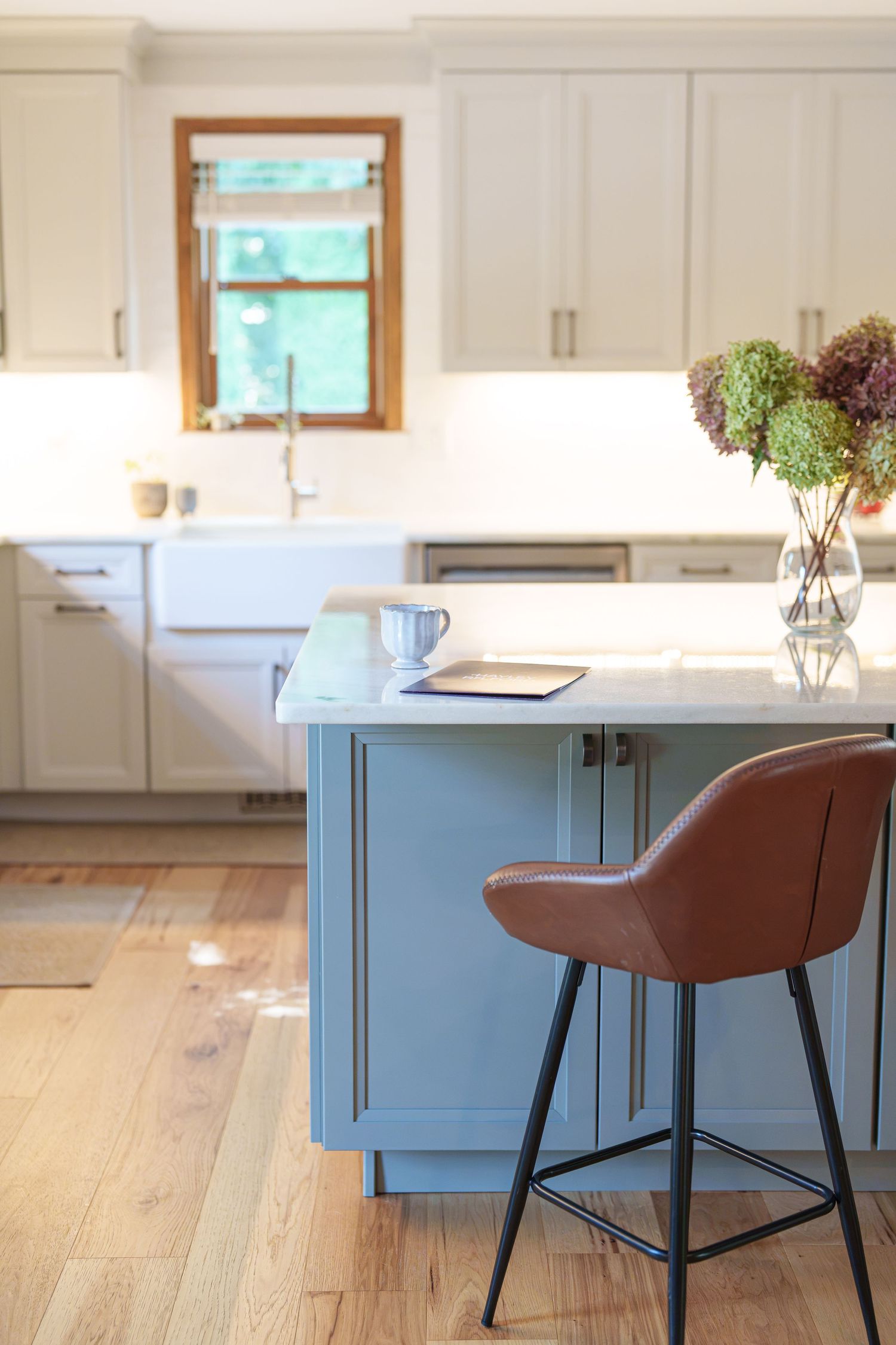 Modern kitchen with blue island, brown leather bar stool, white cabinets, marble countertop, and fresh flowers.