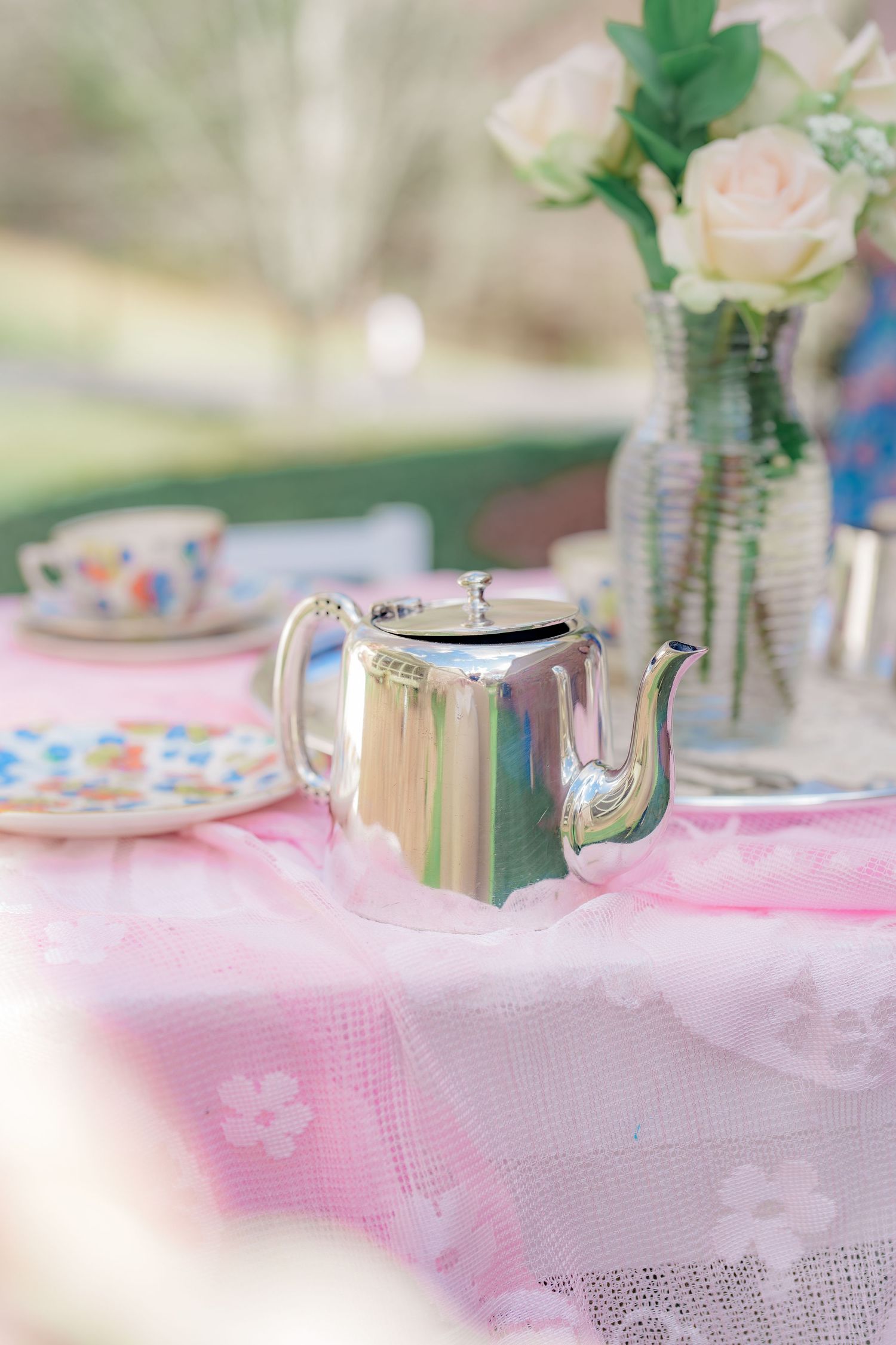 Shiny silver teapot on pink lace tablecloth with colorful teacup and roses at outdoor tea party.