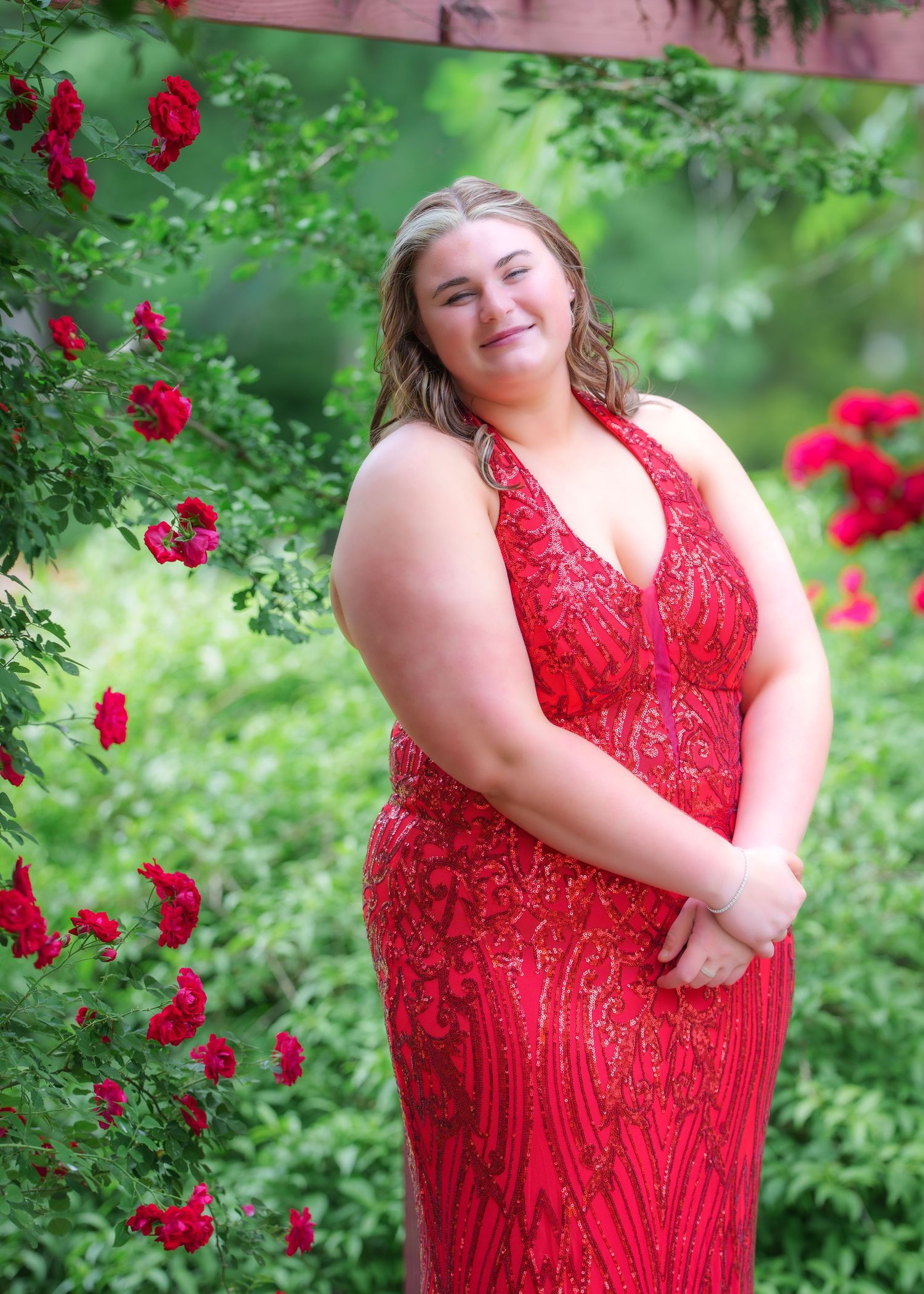 Young woman in a red beaded halter prom gown posing beside blooming red roses in a garden setting.