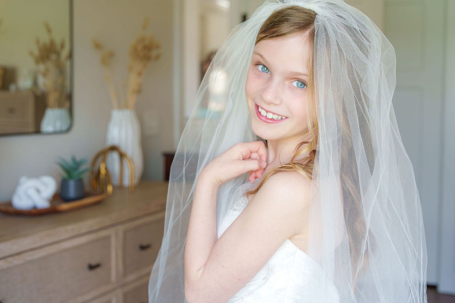 Smiling young girl wearing a white bridal veil poses indoors near a wooden dresser.