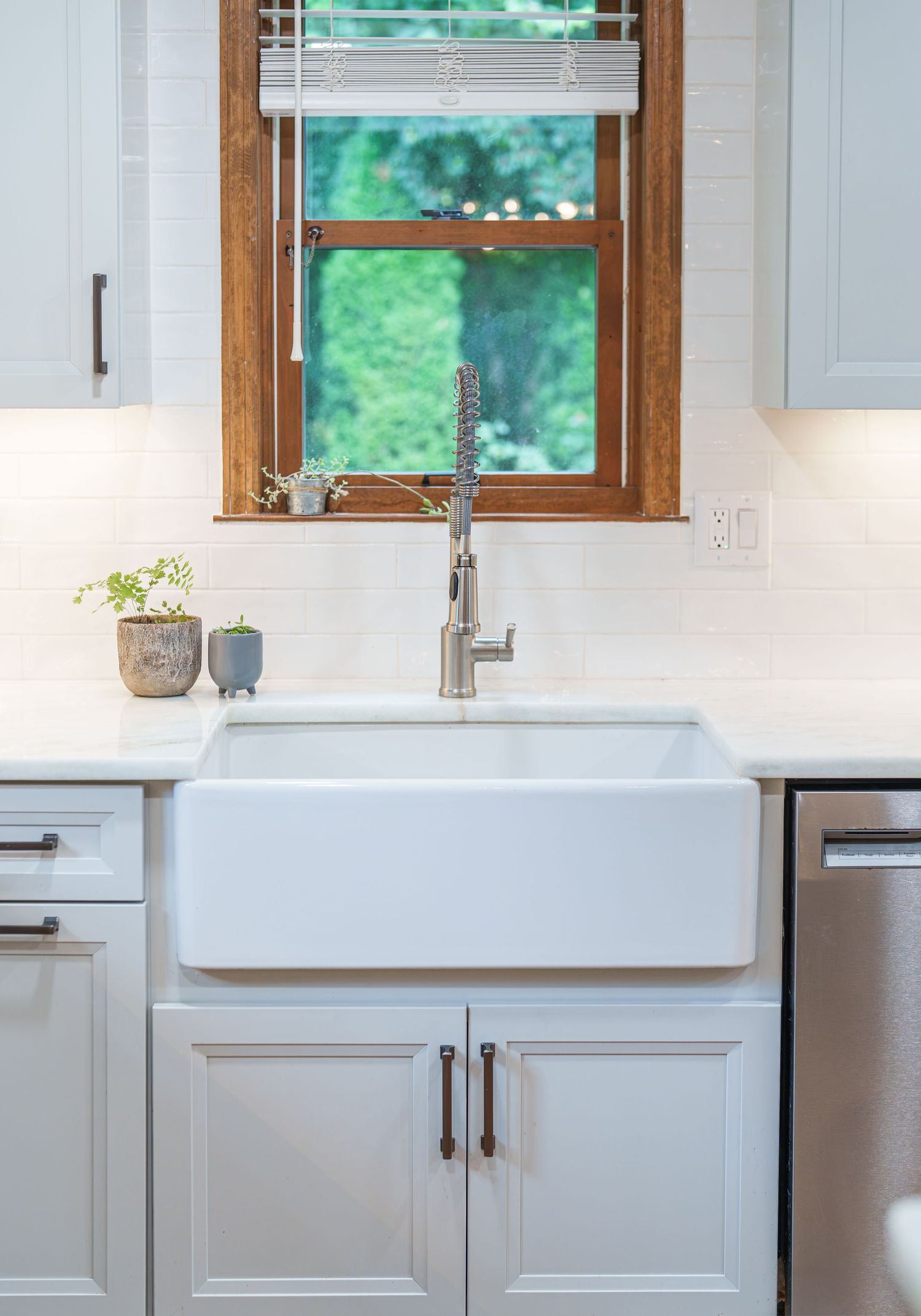 White farmhouse kitchen sink with chrome faucet, gray shaker cabinets, white subway tile backsplash, and wood-framed window.