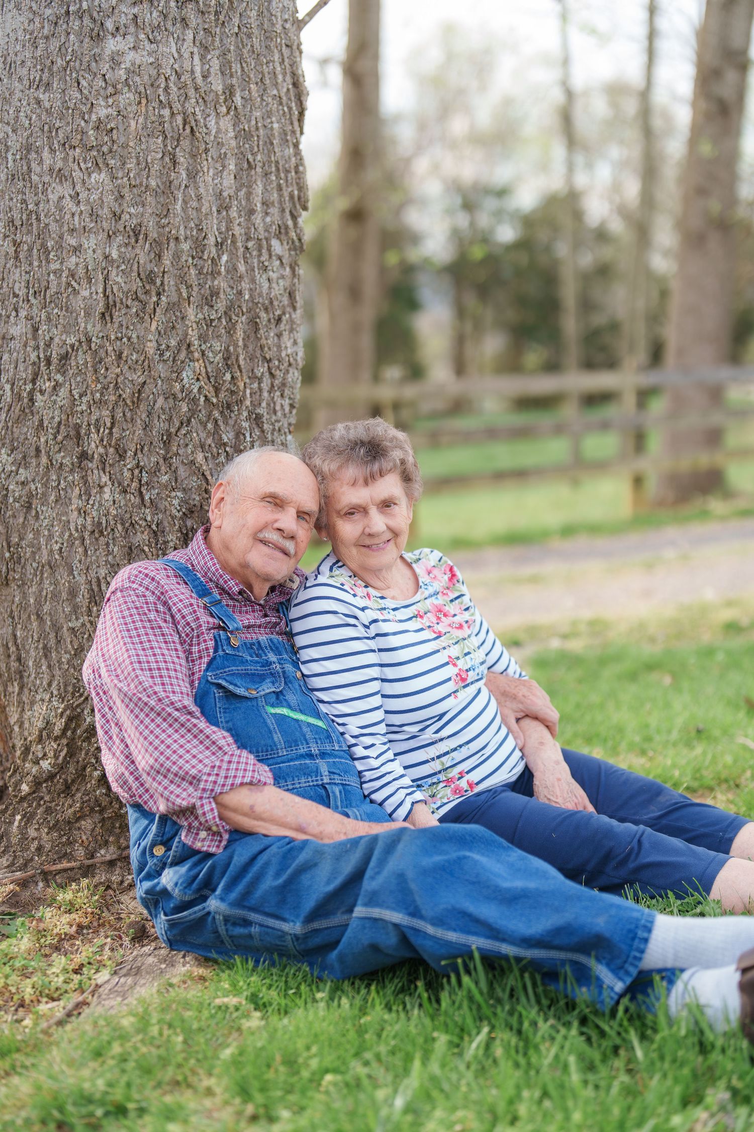 Smiling elderly couple leaning against a tree trunk while sitting on green grass outdoors.