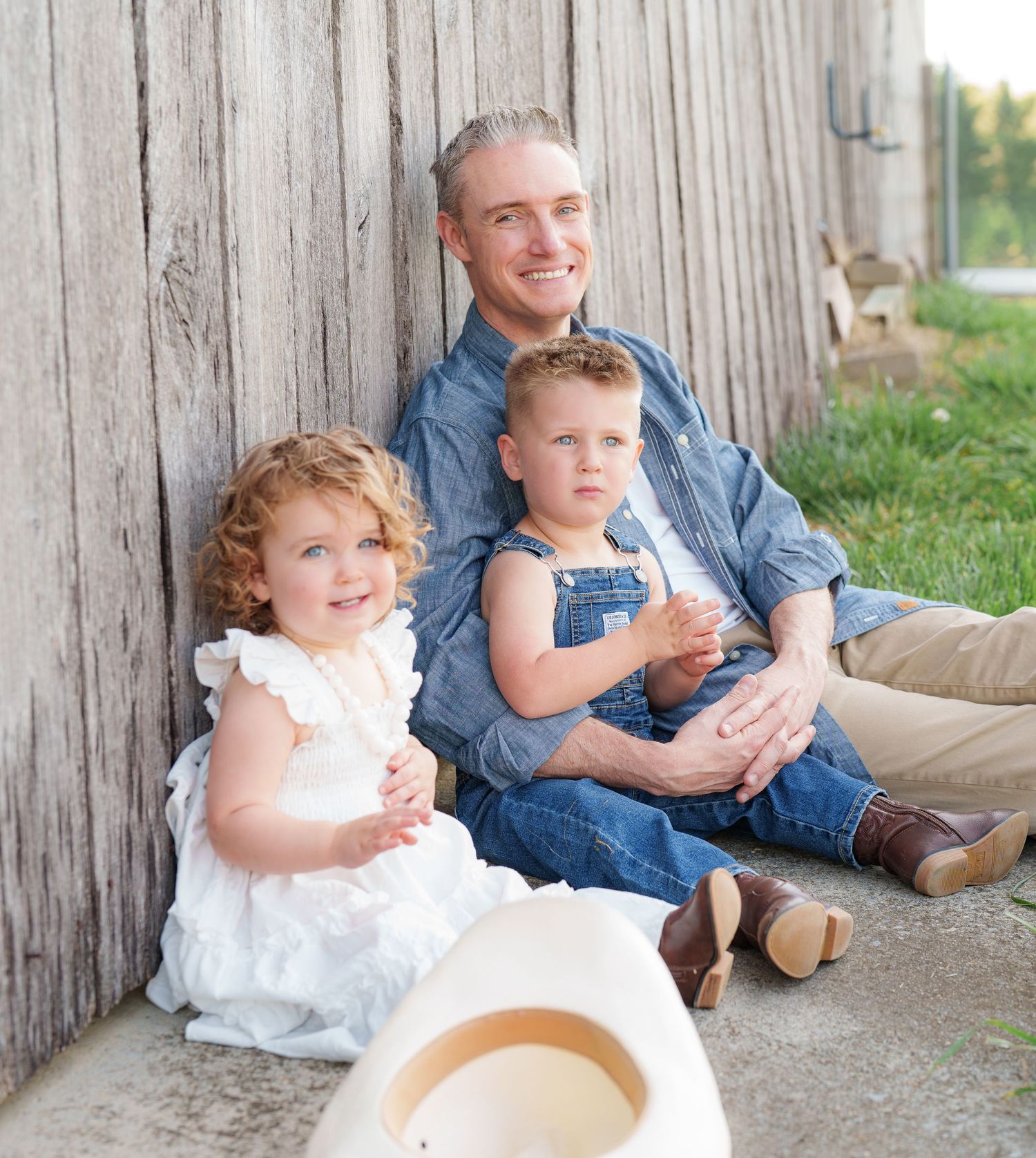 Father sitting against wooden fence with two young children in denim overalls outdoors.