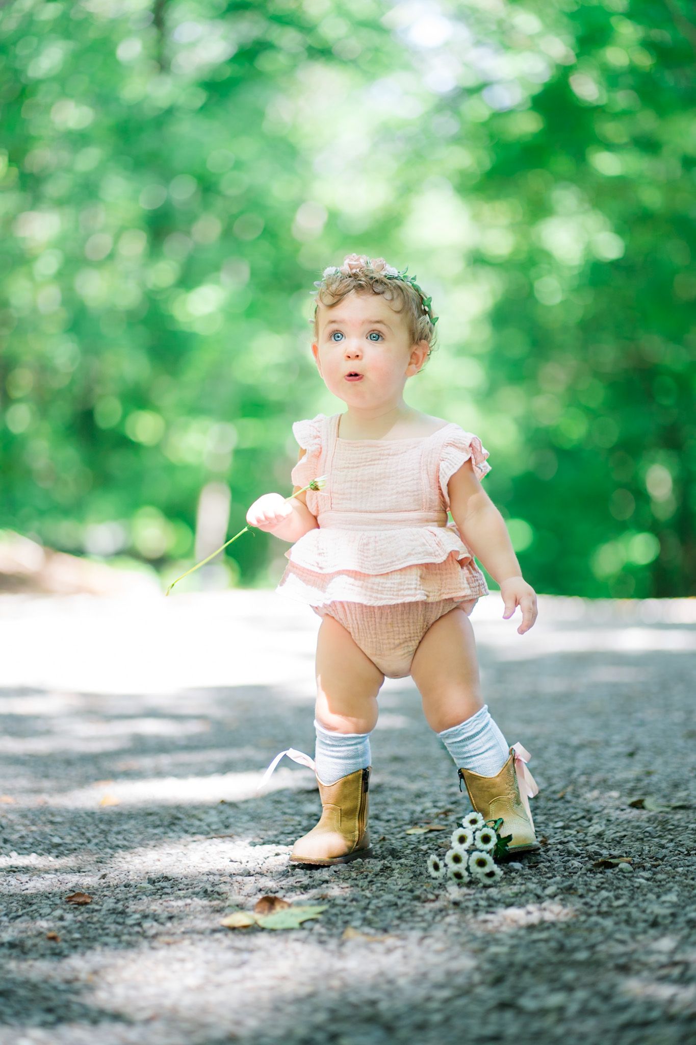 Adorable baby girl in pink romper, floral crown, and gold boots standing on a path surrounded by lush green trees.