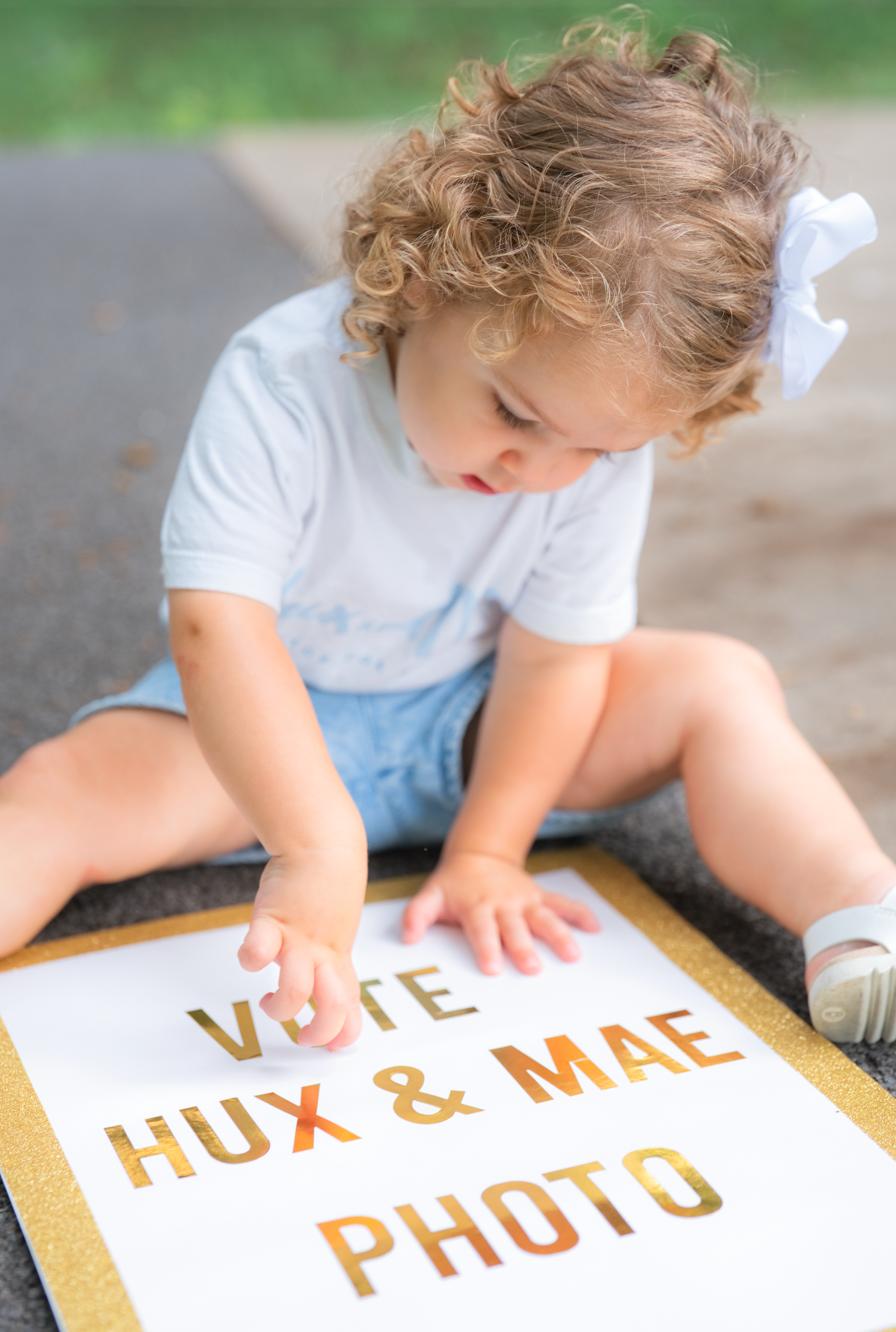 Curly-haired toddler sitting on pavement holding a glittery sign reading Vote Hux & Mae Photo.