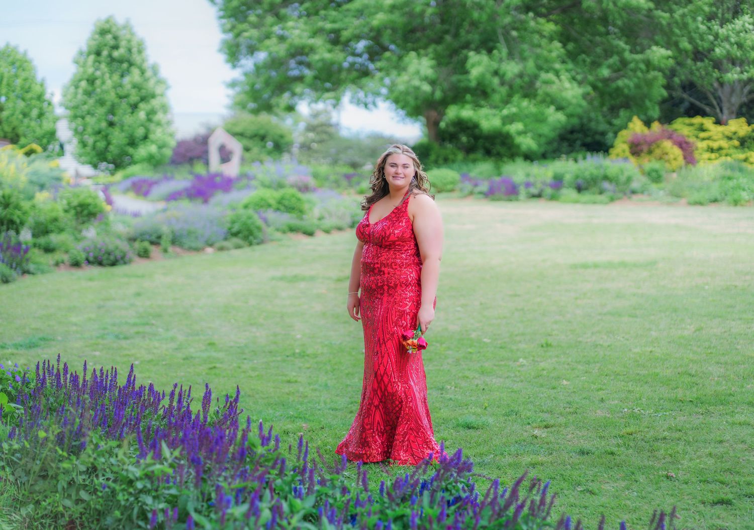 Young woman in red lace gown poses in lush garden with purple flowers during prom or formal event.