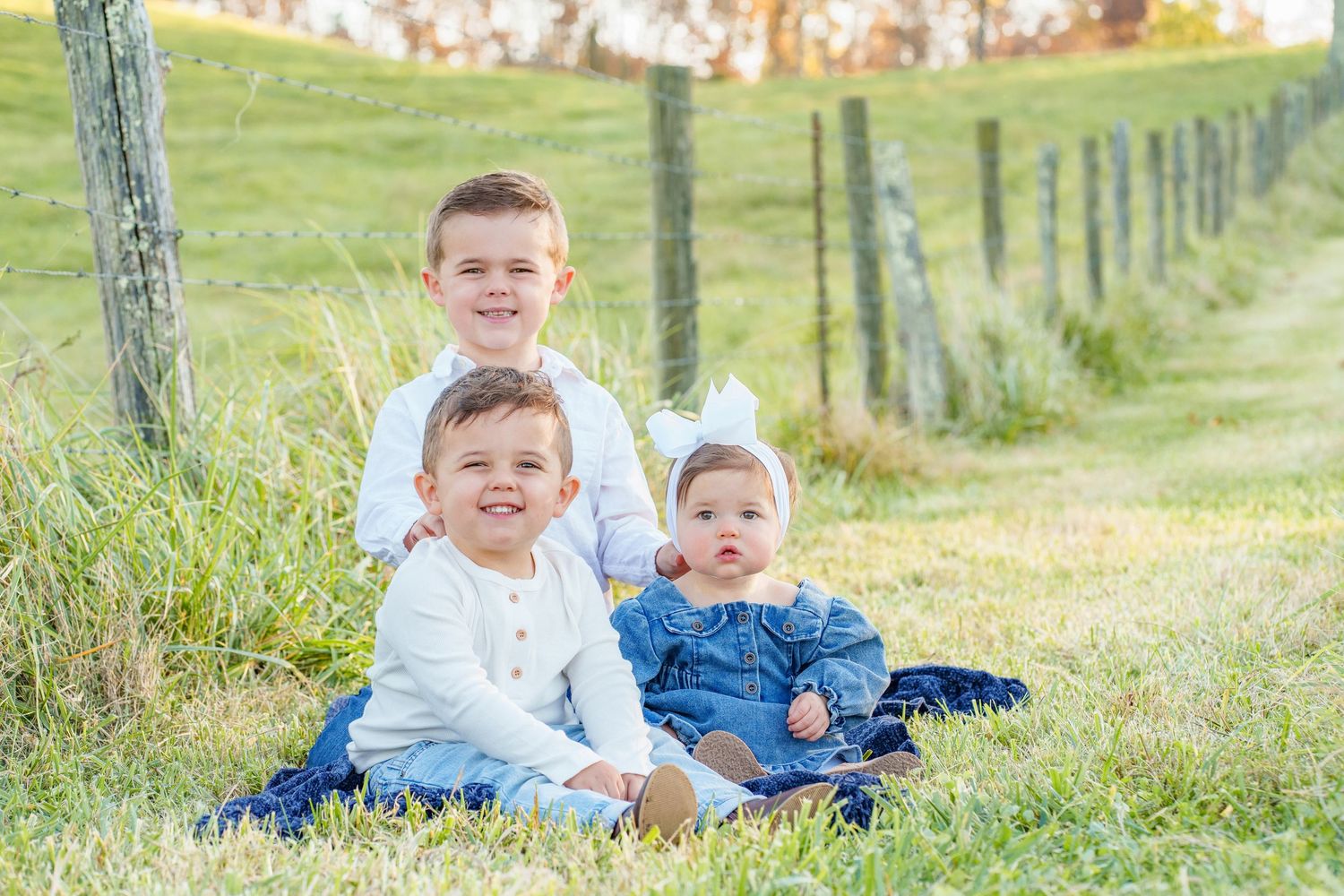 Three young siblings smile together while sitting on grass beside a wooden fence in a sunny outdoor setting.