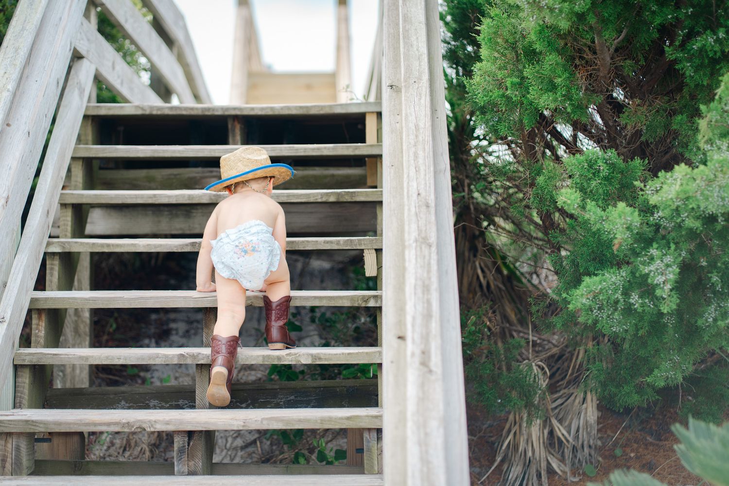 Toddler in cowboy hat and boots climbs wooden outdoor stairs near green tree.