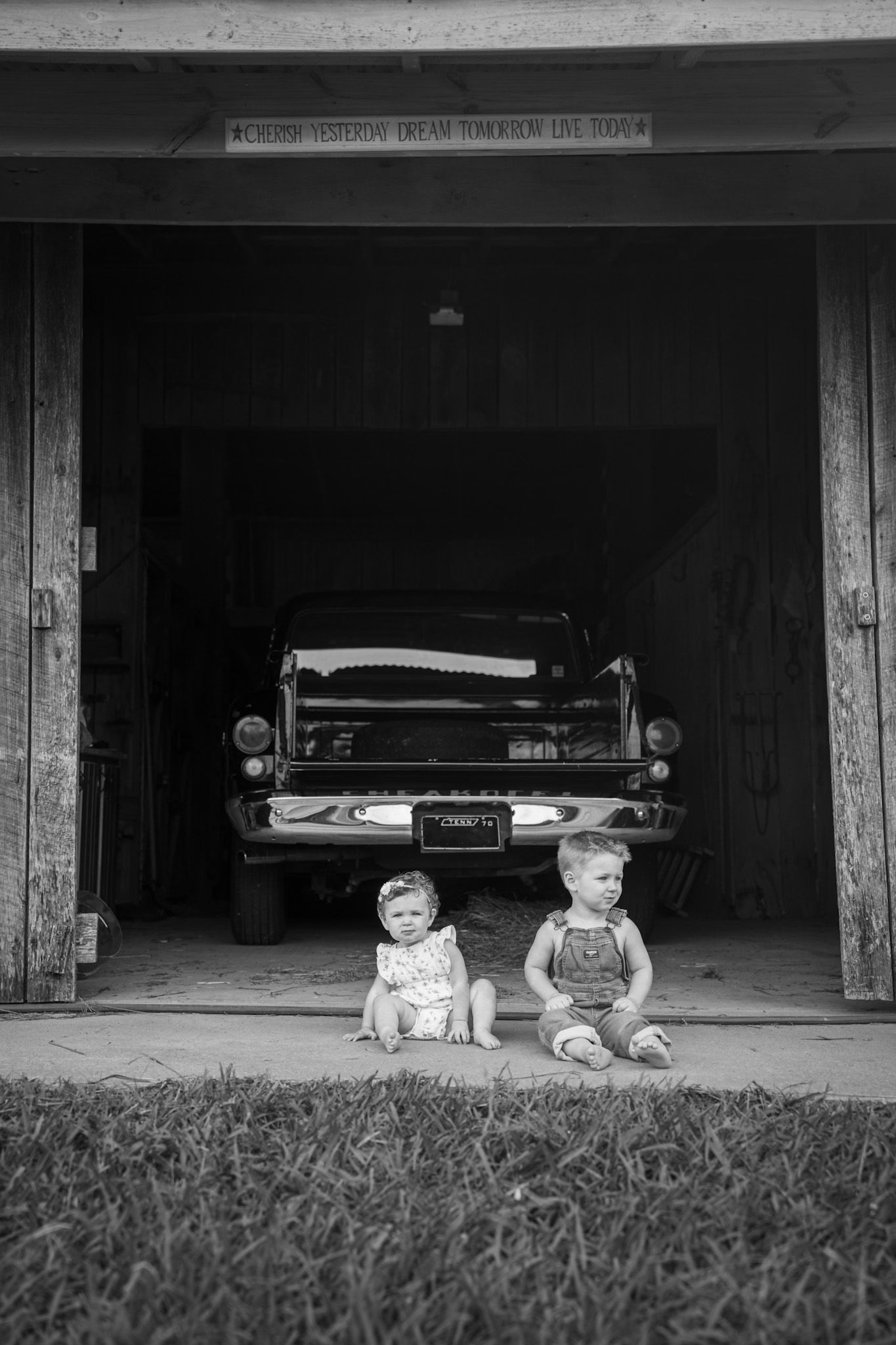 Two young siblings sit in garage doorway in front of classic vintage black car.