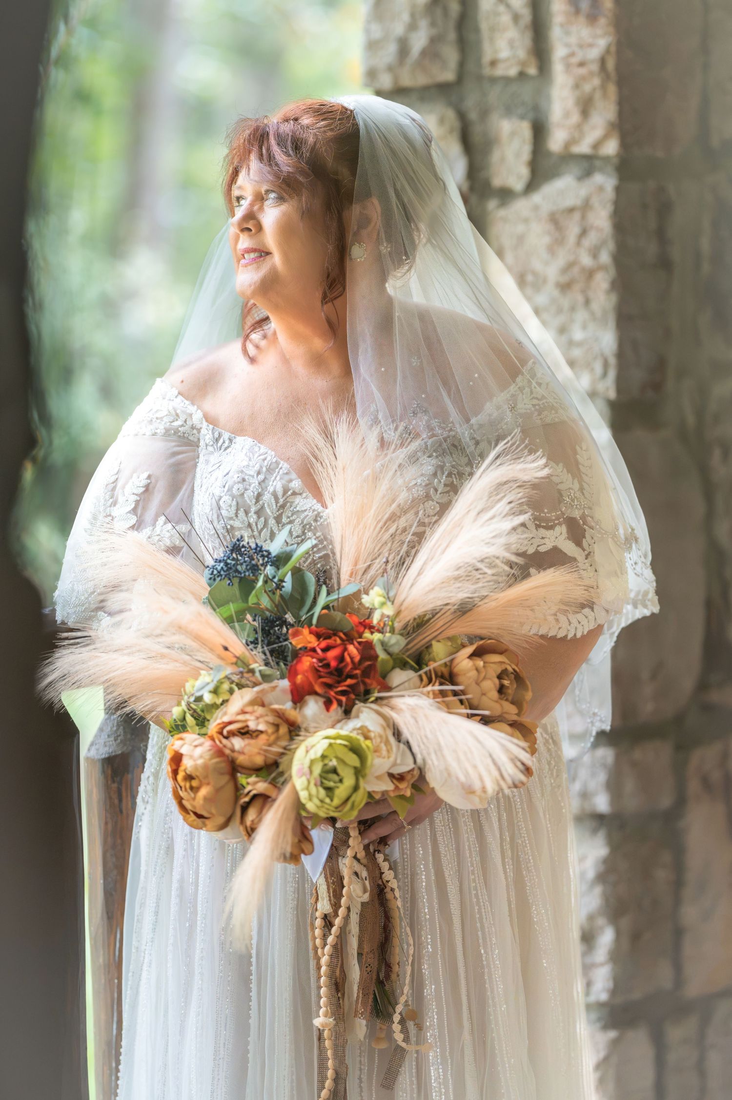 Bride in white lace gown and veil holds boho bouquet with pampas grass and roses near stone column in Gatlinburg, TN.