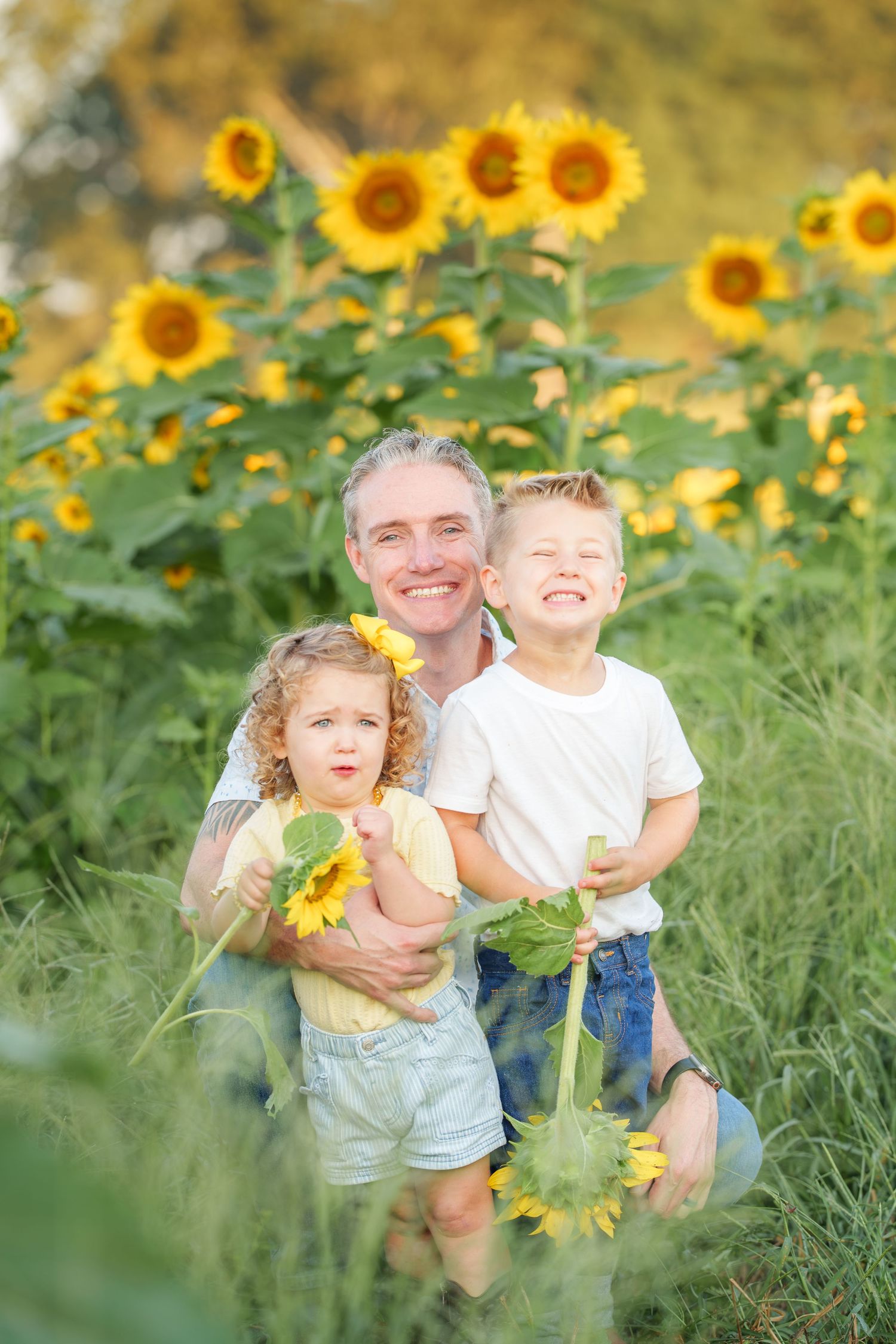 Father kneeling with two young children holding sunflowers in a vibrant sunflower field on a sunny day.