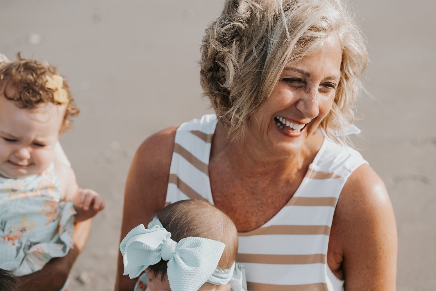 Smiling grandmother holding two babies at the beach on a sunny day.