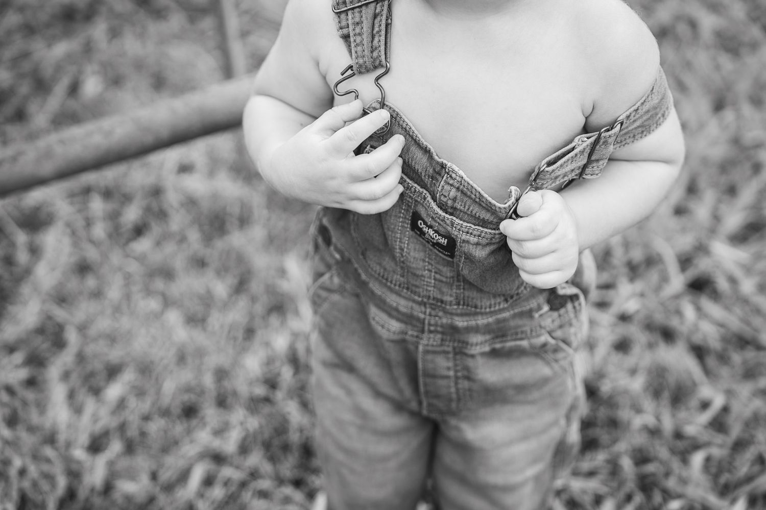 Close-up of toddler hands gripping denim overall straps in grassy outdoor setting.