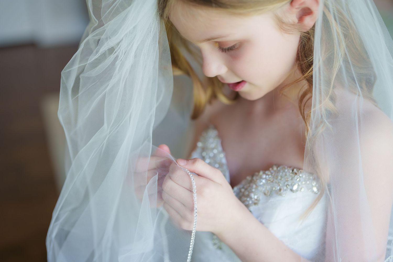 Young flower girl in white beaded dress and veil carefully holding delicate ribbon at a wedding ceremony.