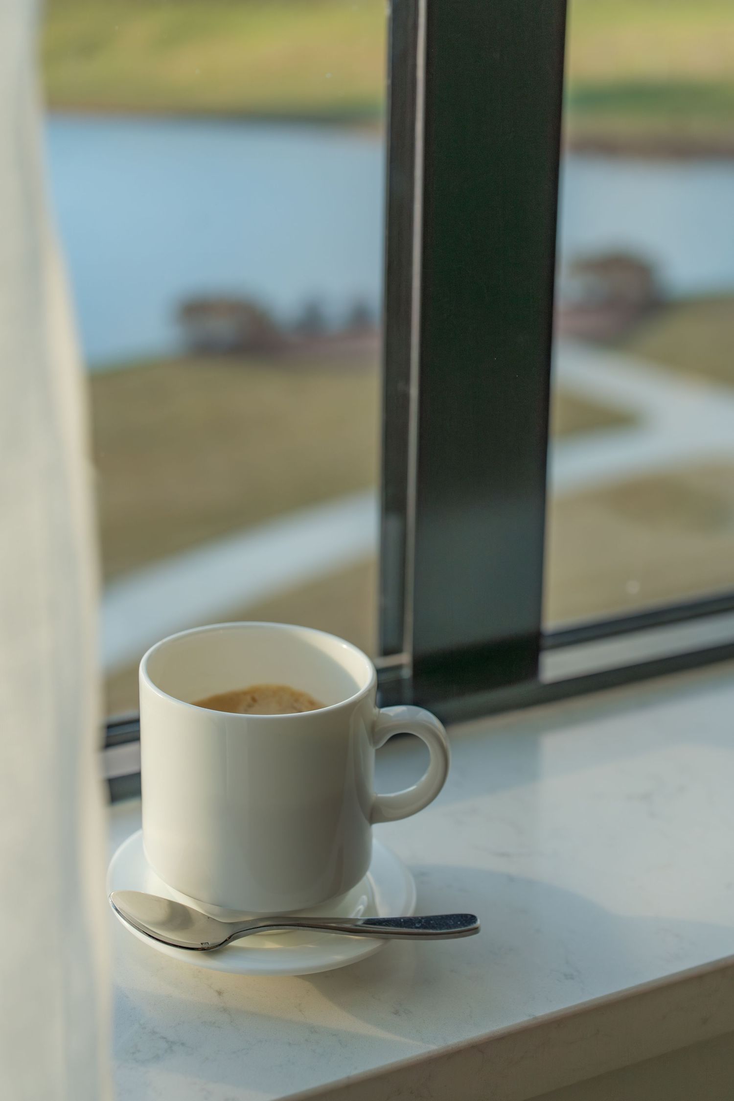 White coffee cup on saucer with spoon on marble windowsill overlooking scenic waterway and green landscape.