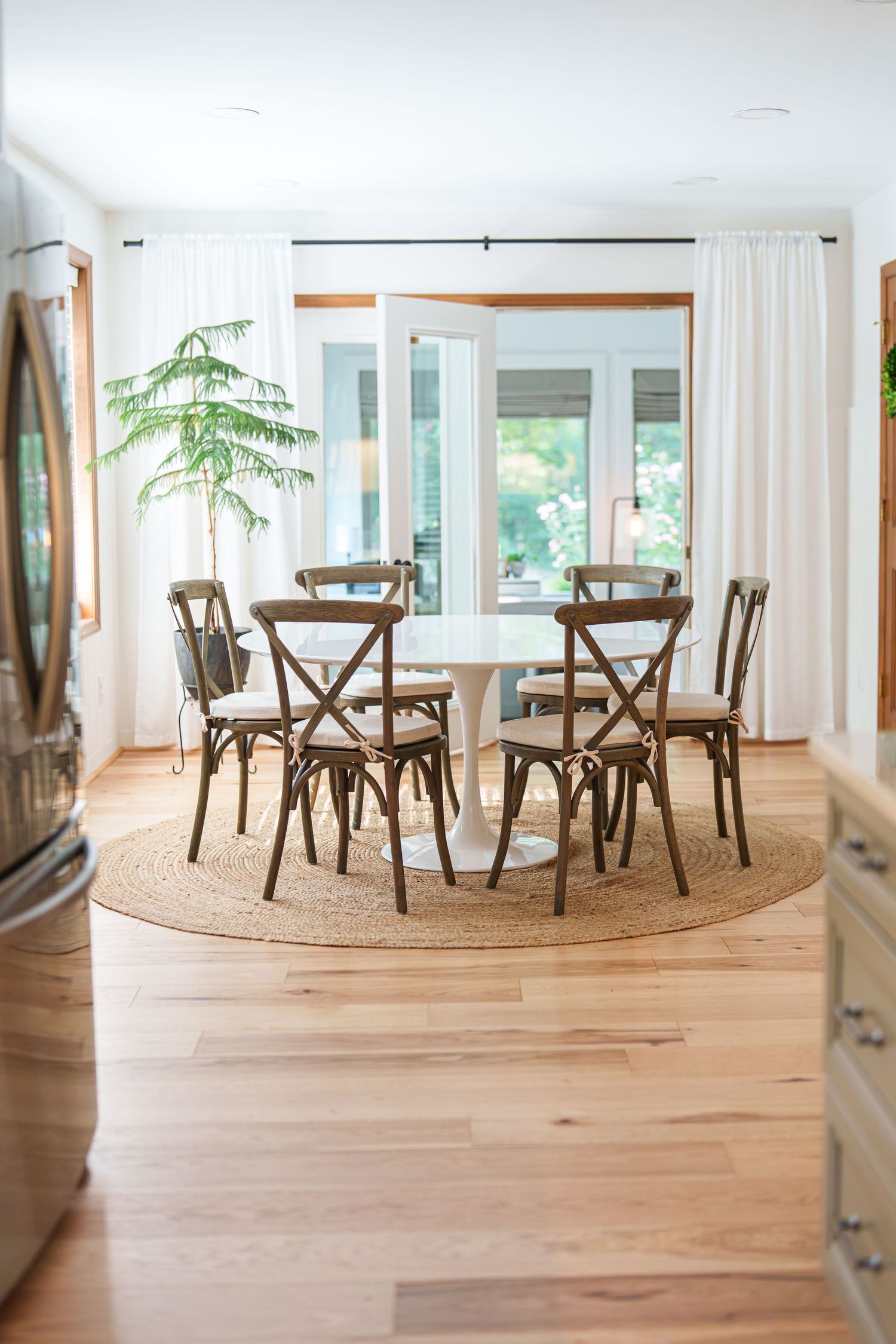 Bright dining room with white round table, cross-back chairs on jute rug, hardwood floors, and large windows with white curtains.