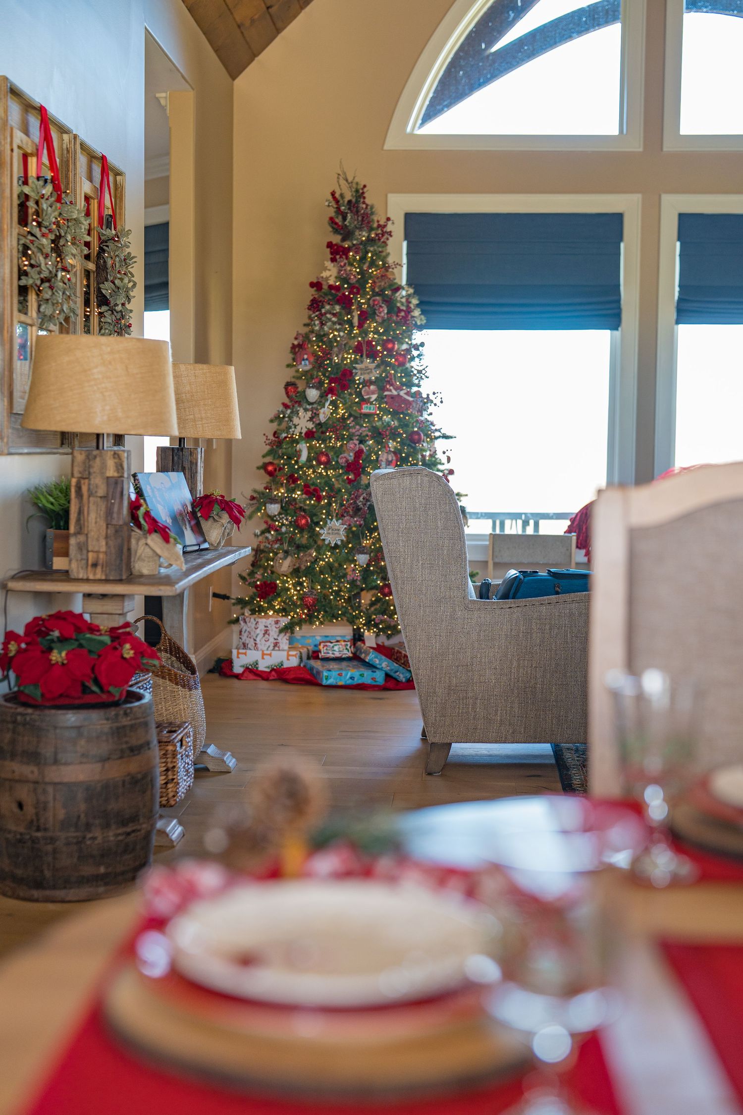 Festive living room with decorated Christmas tree, wrapped gifts, red table settings, and large arched windows in a cozy home.