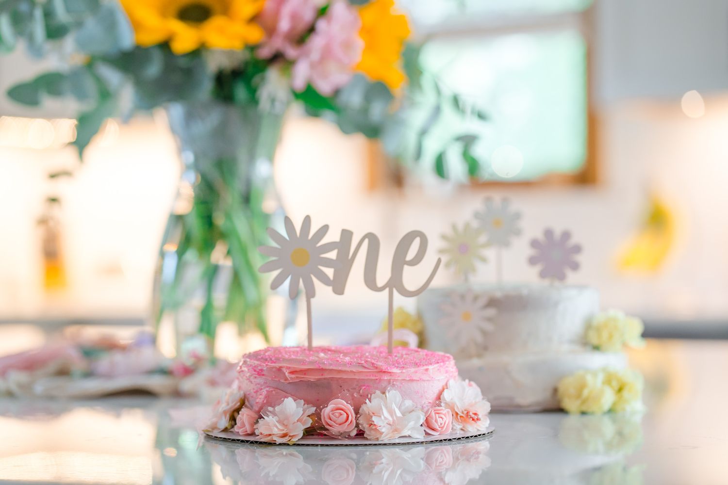 Pink first birthday smash cake with floral decorations and a daisy 'one' topper on a kitchen counter.
