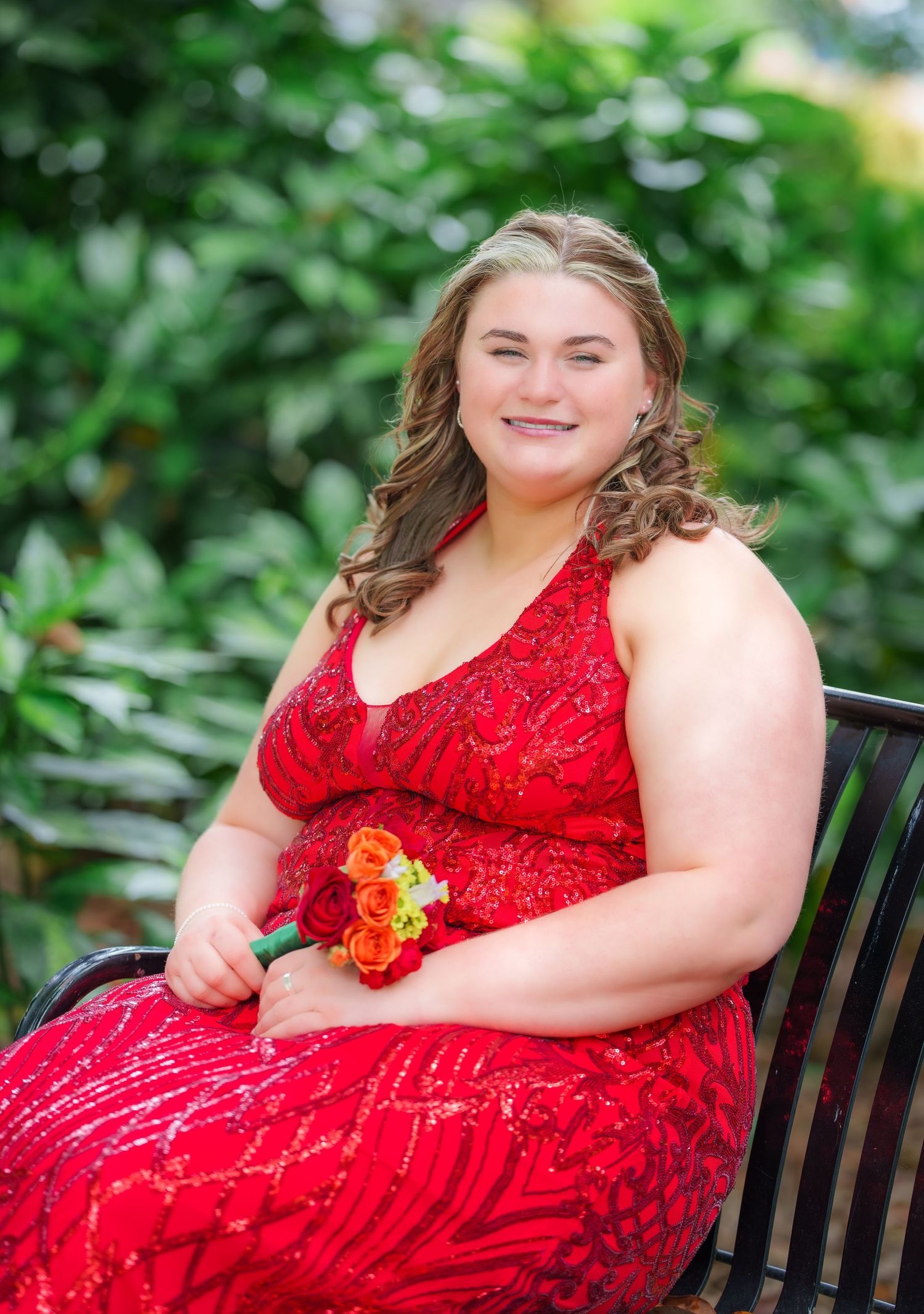Smiling young woman in red sequin gown holding orange roses while seated on a bench.