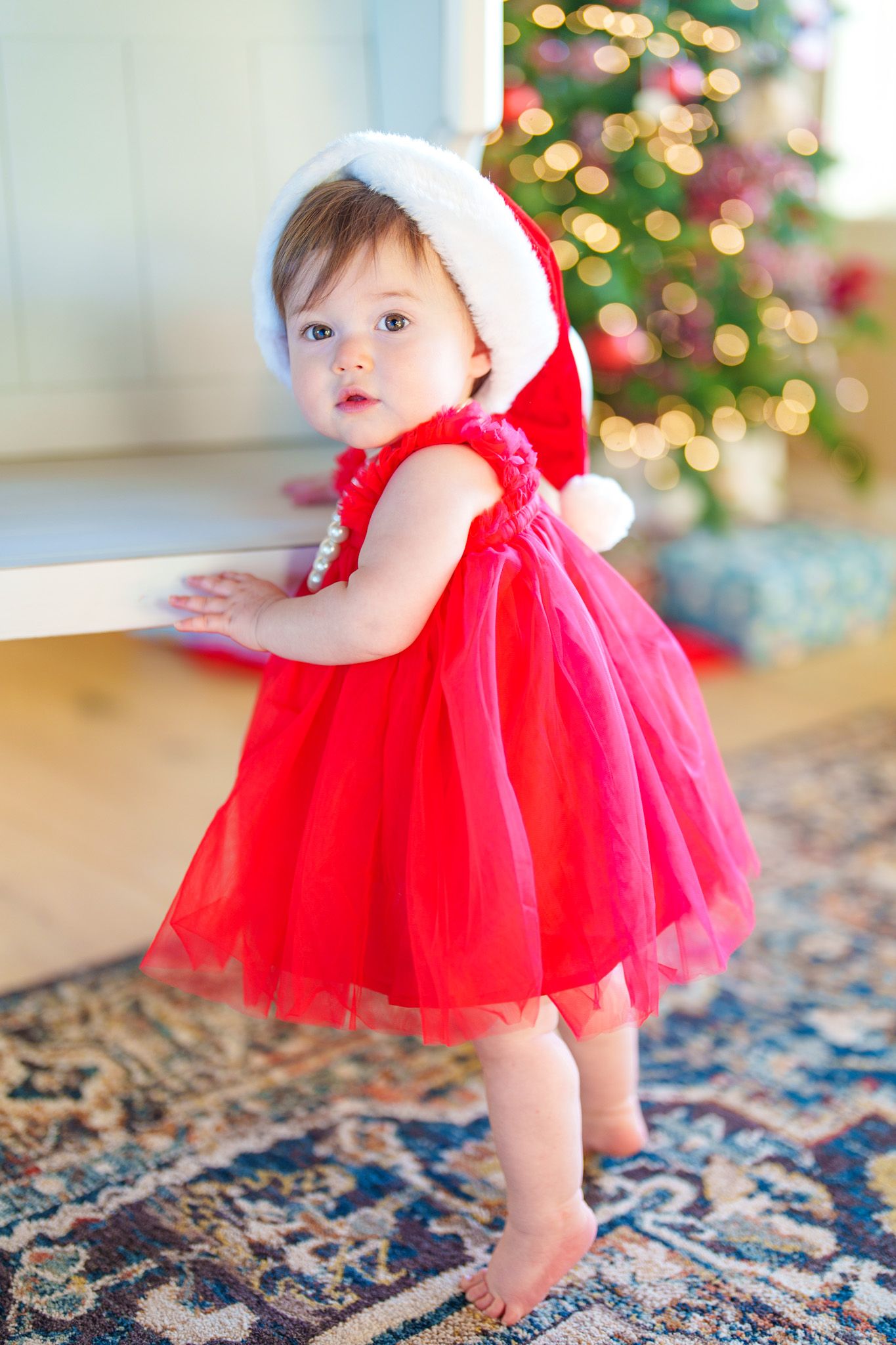 Baby girl in red tutu dress and Santa hat standing barefoot near a decorated Christmas tree.