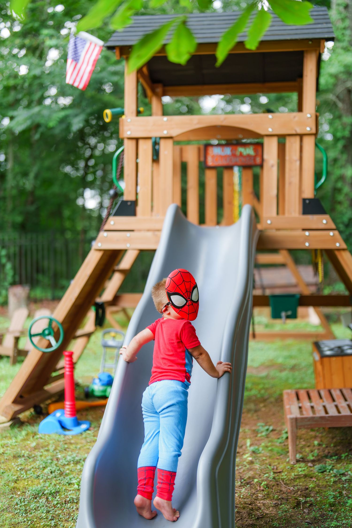 Child in Spiderman costume climbing a backyard wooden playground slide on a sunny day.