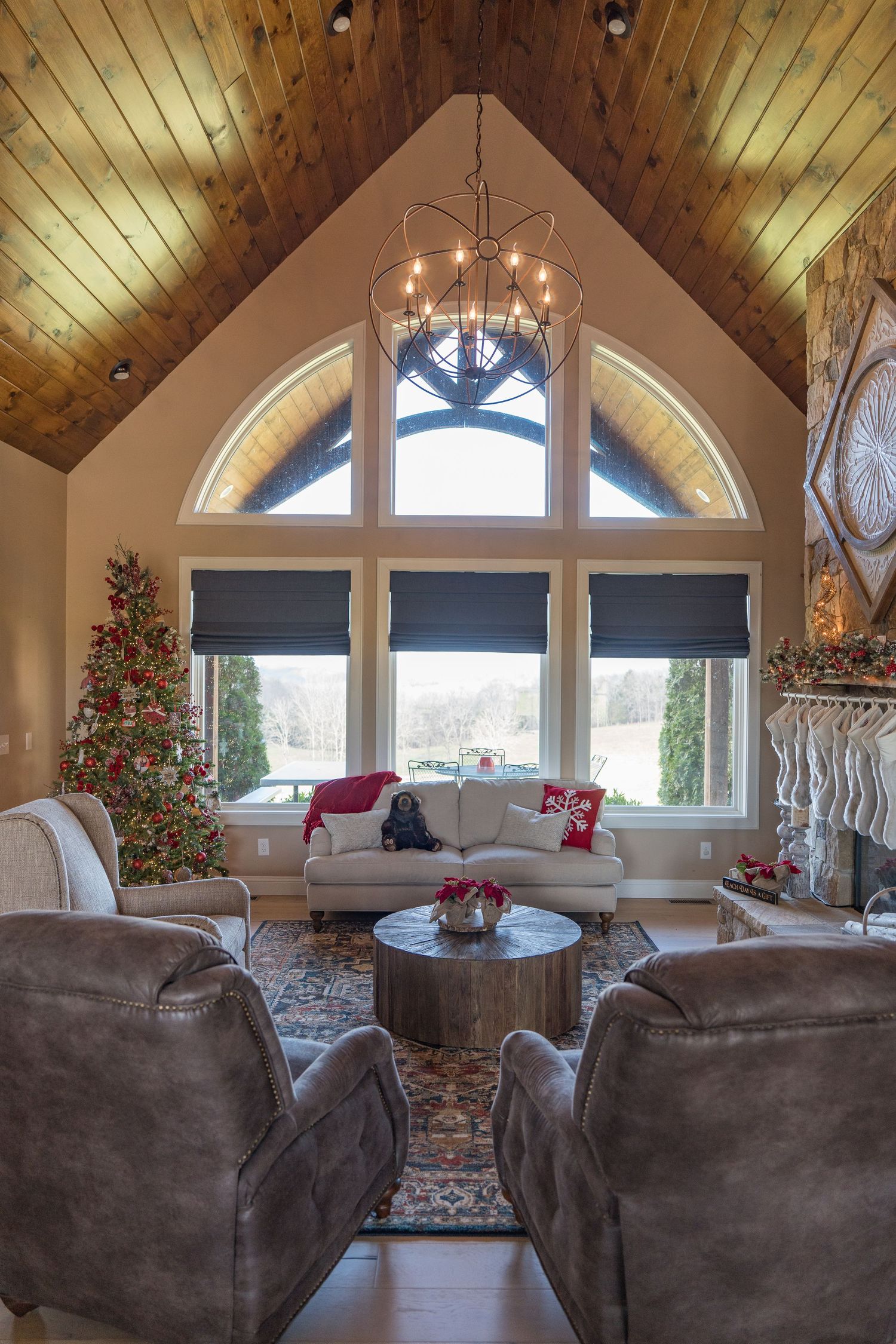 Cozy living room with vaulted wood ceiling, chandelier, Christmas tree, fireplace, and large arched windows overlooking snowy landscape.