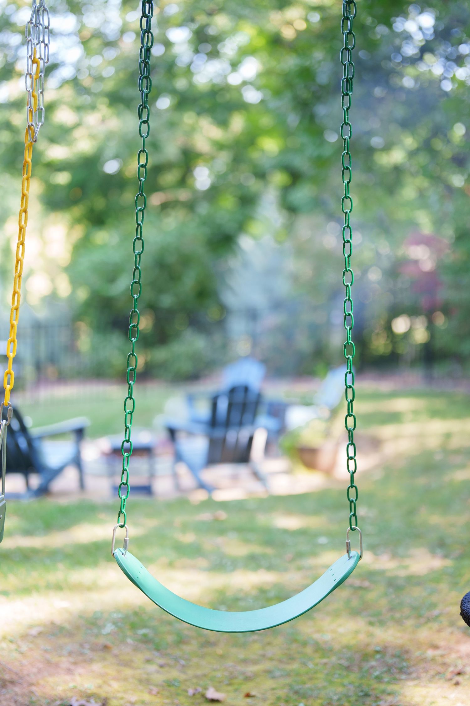 Green belt swing with chain hanging in a backyard with Adirondack chairs and fire pit in background.