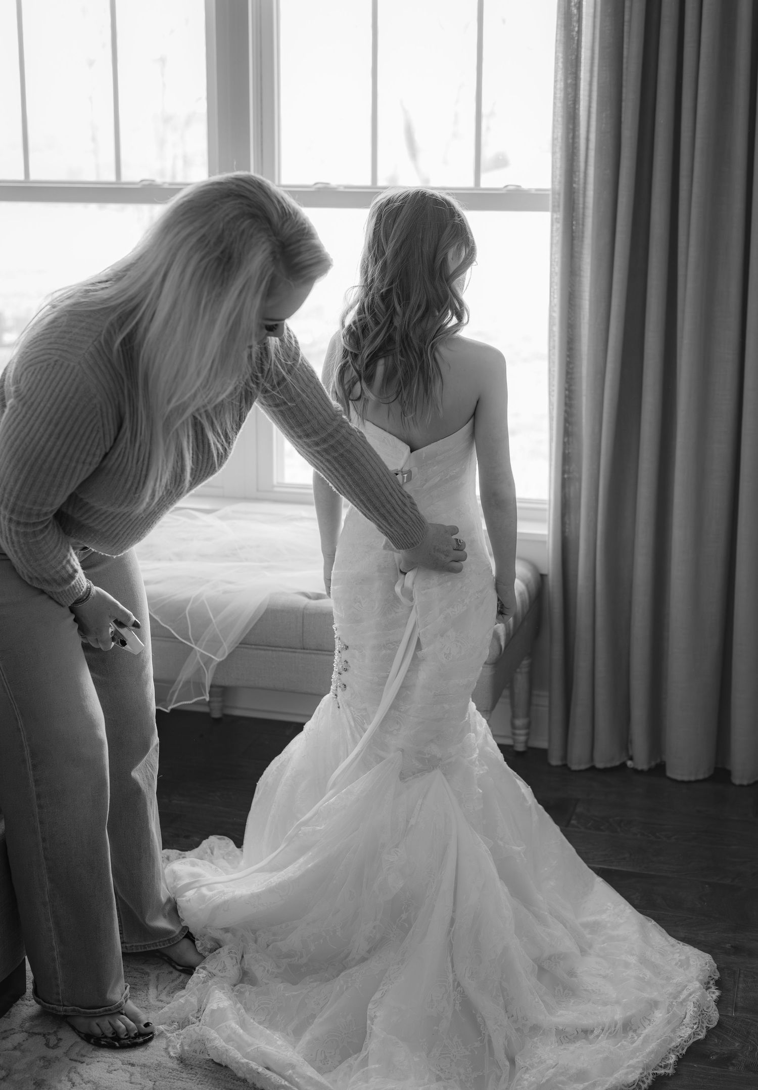 Black and white photo of a woman helping a bride in a lace mermaid wedding dress near a window.