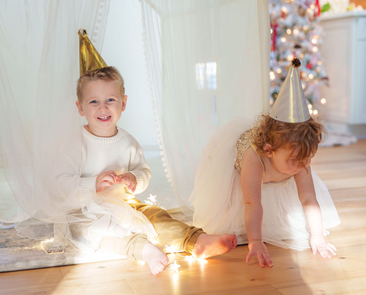 Two young children in party hats playing with string lights near a Christmas tree.
