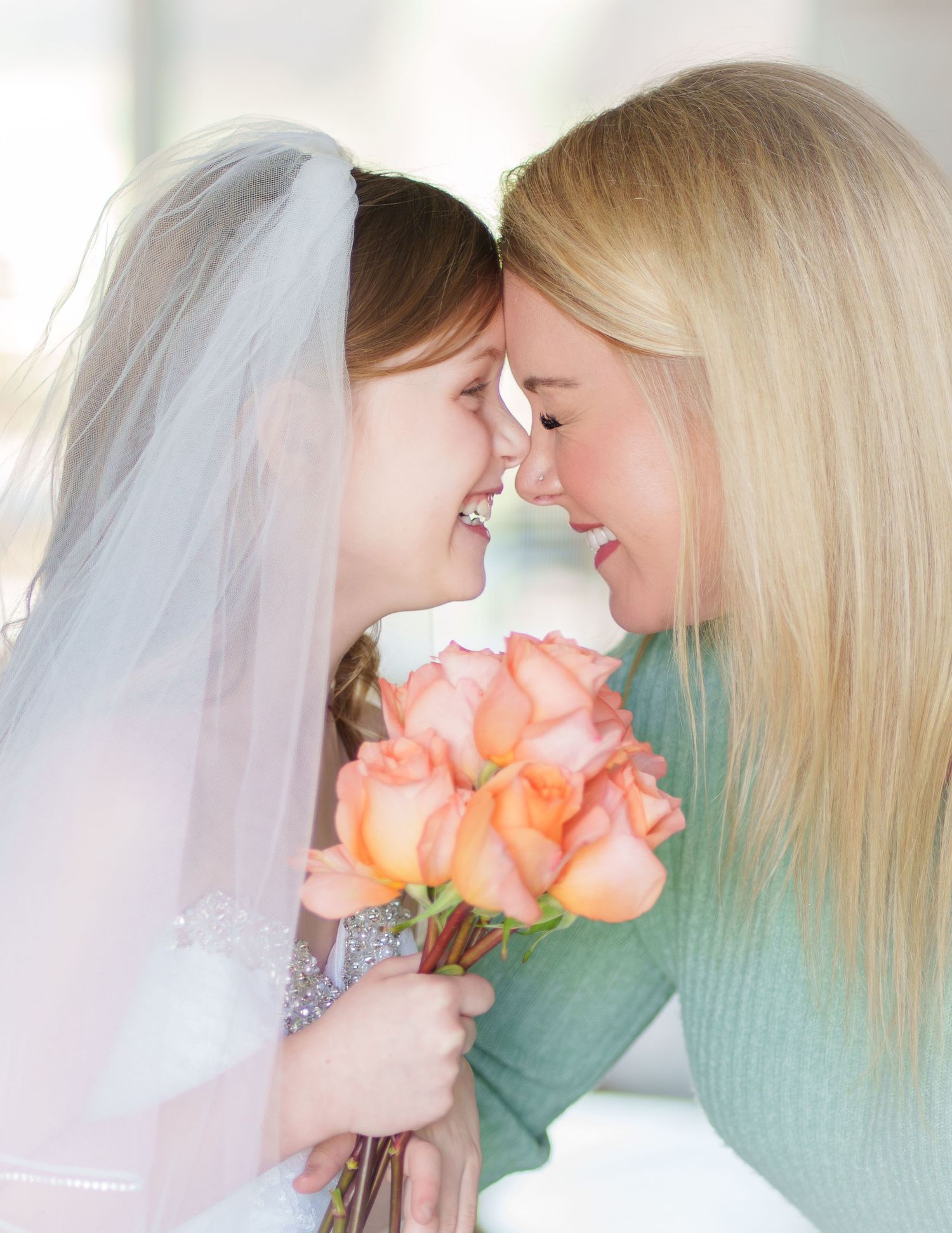 Young flower girl in veil holds peach roses, touching foreheads with smiling blonde woman in tender moment.