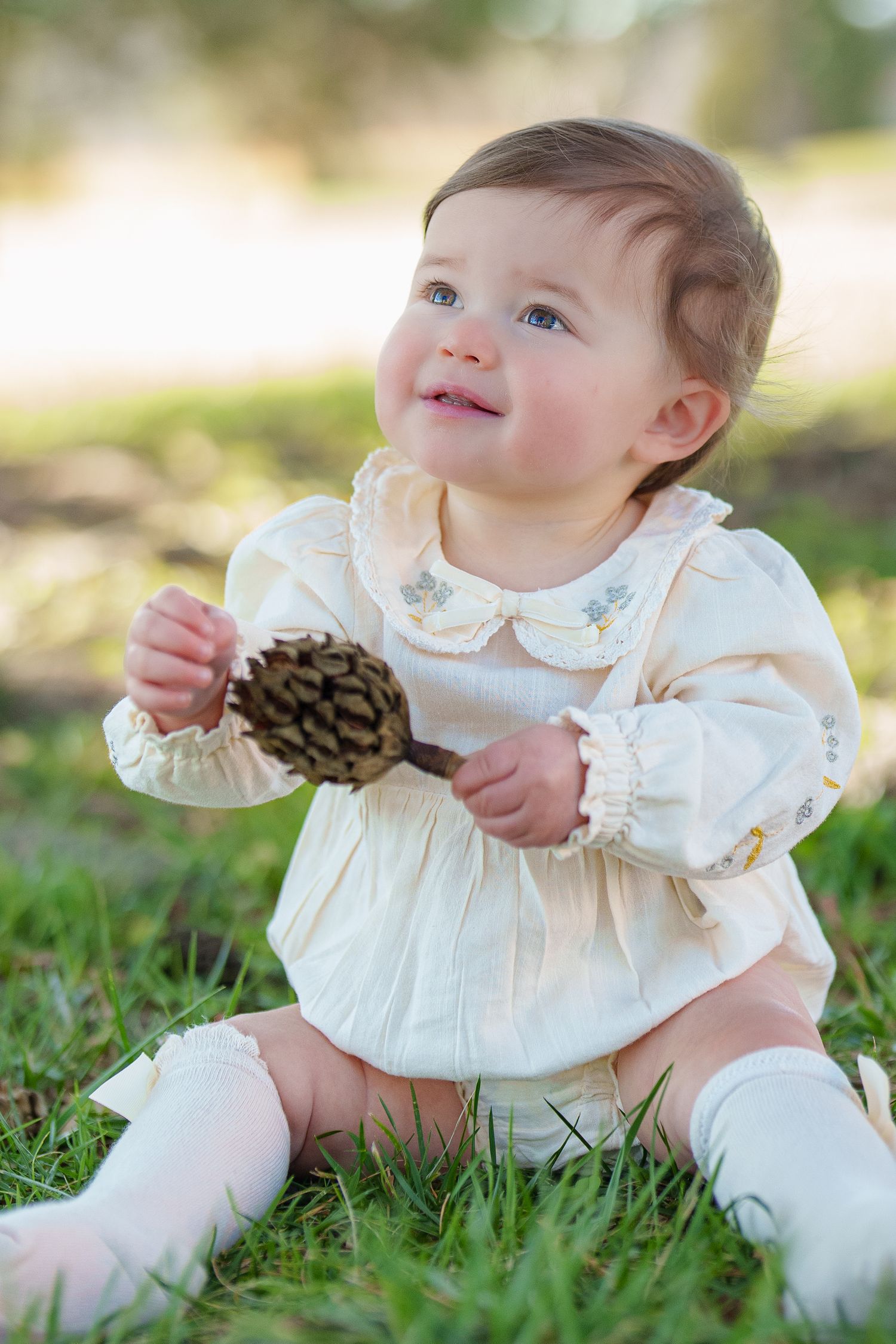 Smiling baby girl in cream dress sitting on grass holding a pine cone outdoors.