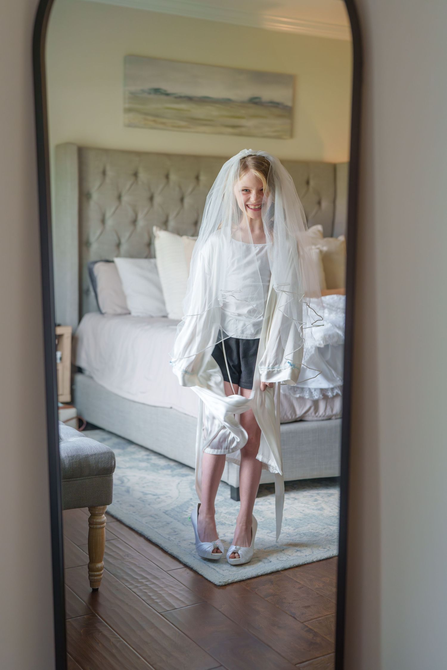 Young girl in wedding veil and oversized heels poses in mirror reflection in bedroom.