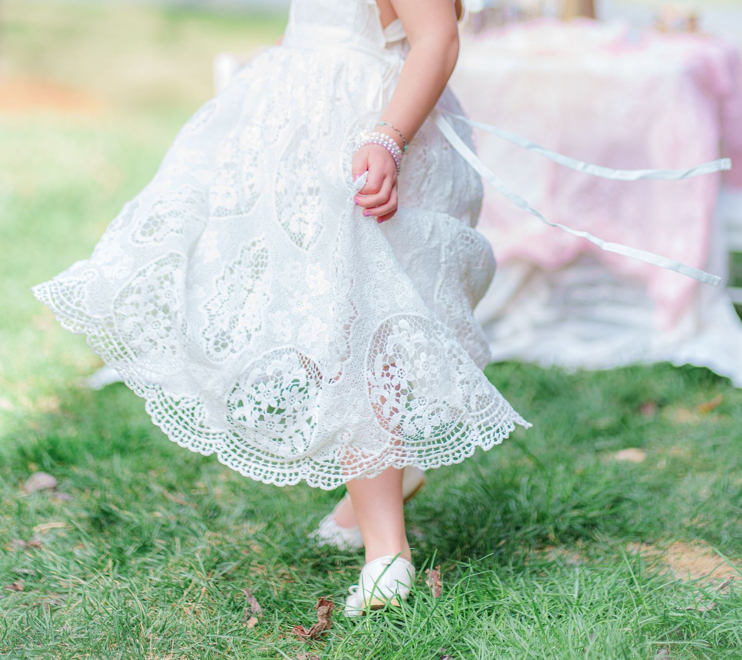 A young girl twirls in a white lace dress on green grass at an outdoor party.