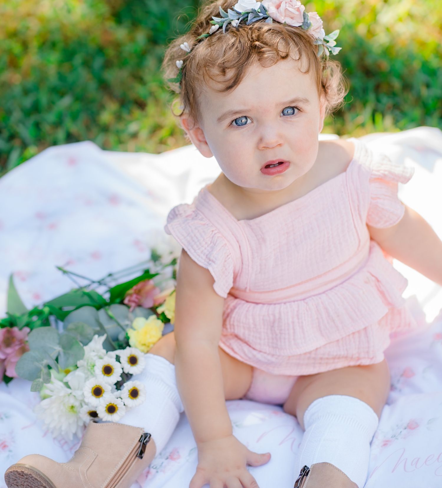 Blue-eyed baby girl in pink romper and floral crown sitting on blanket outdoors with flowers.