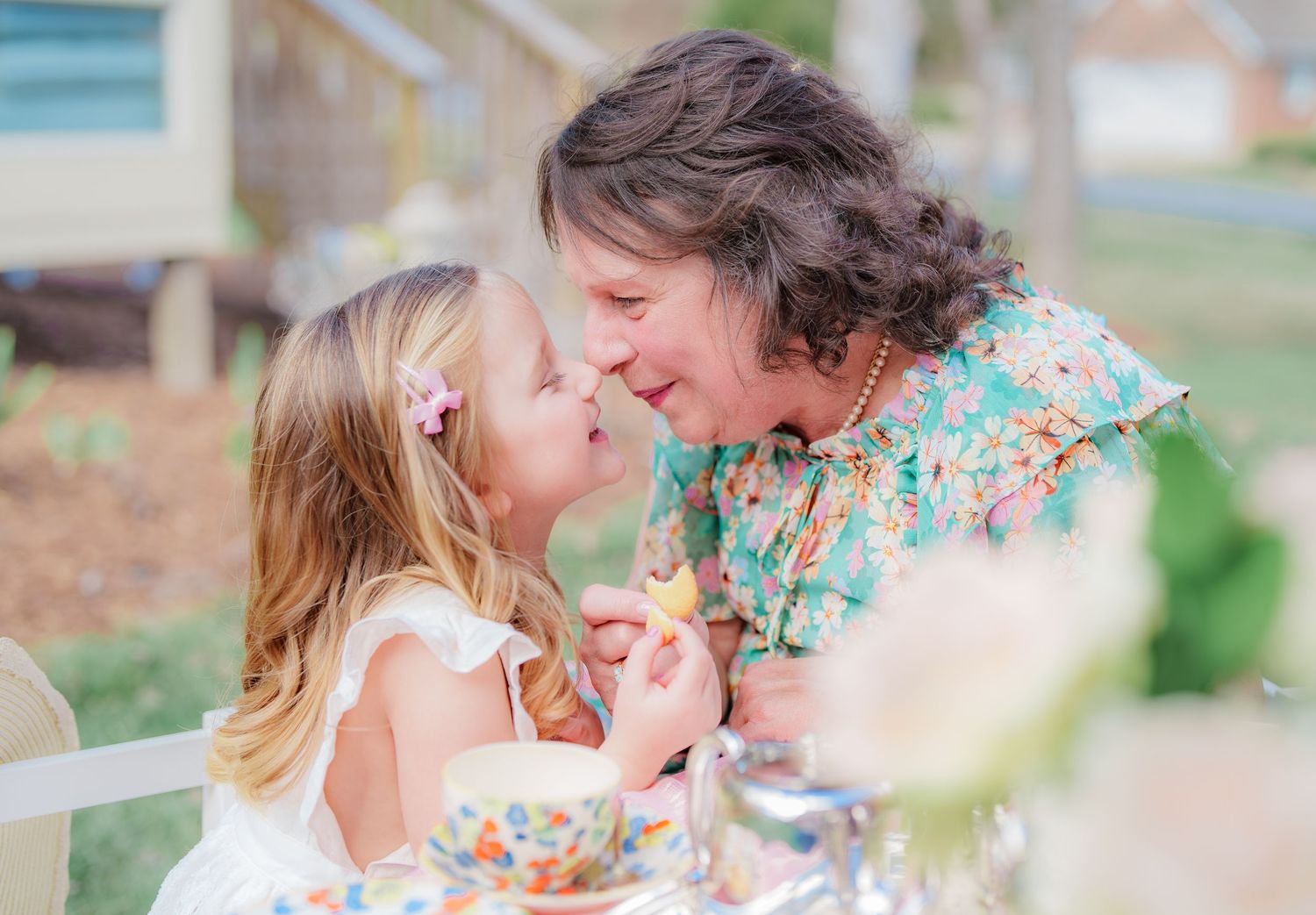 A grandmother and young granddaughter share a sweet nose-to-nose moment at an outdoor tea party table.