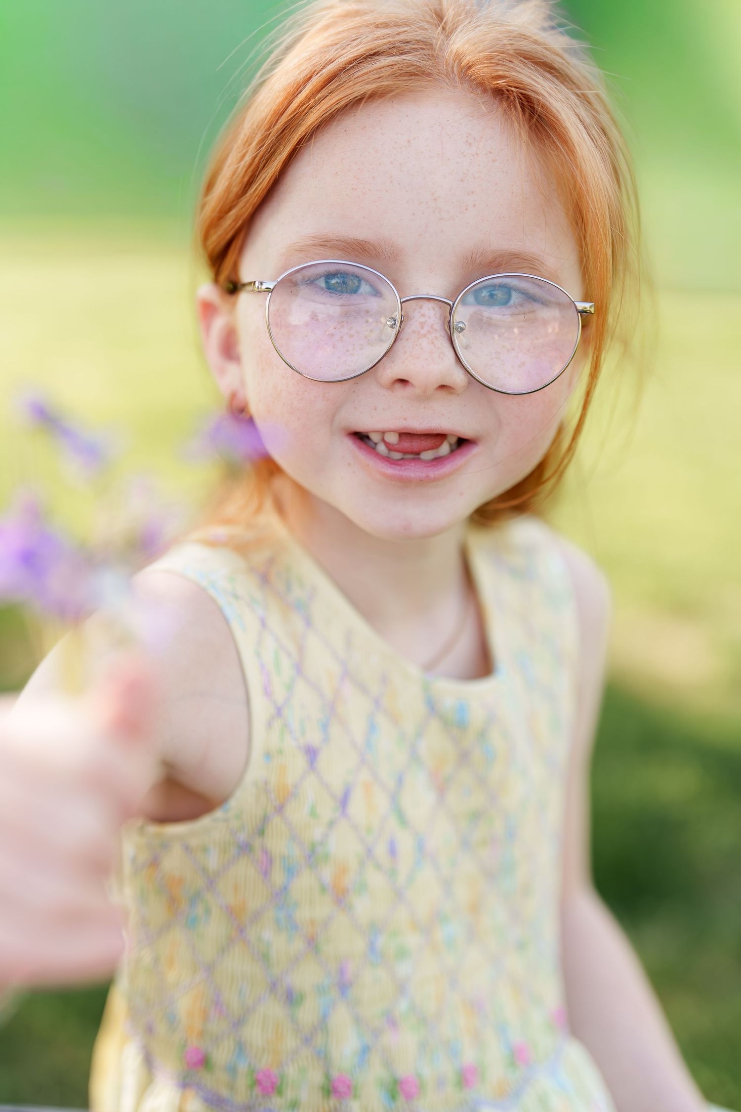 Smiling redhead girl with freckles and round glasses holding purple wildflowers outdoors.