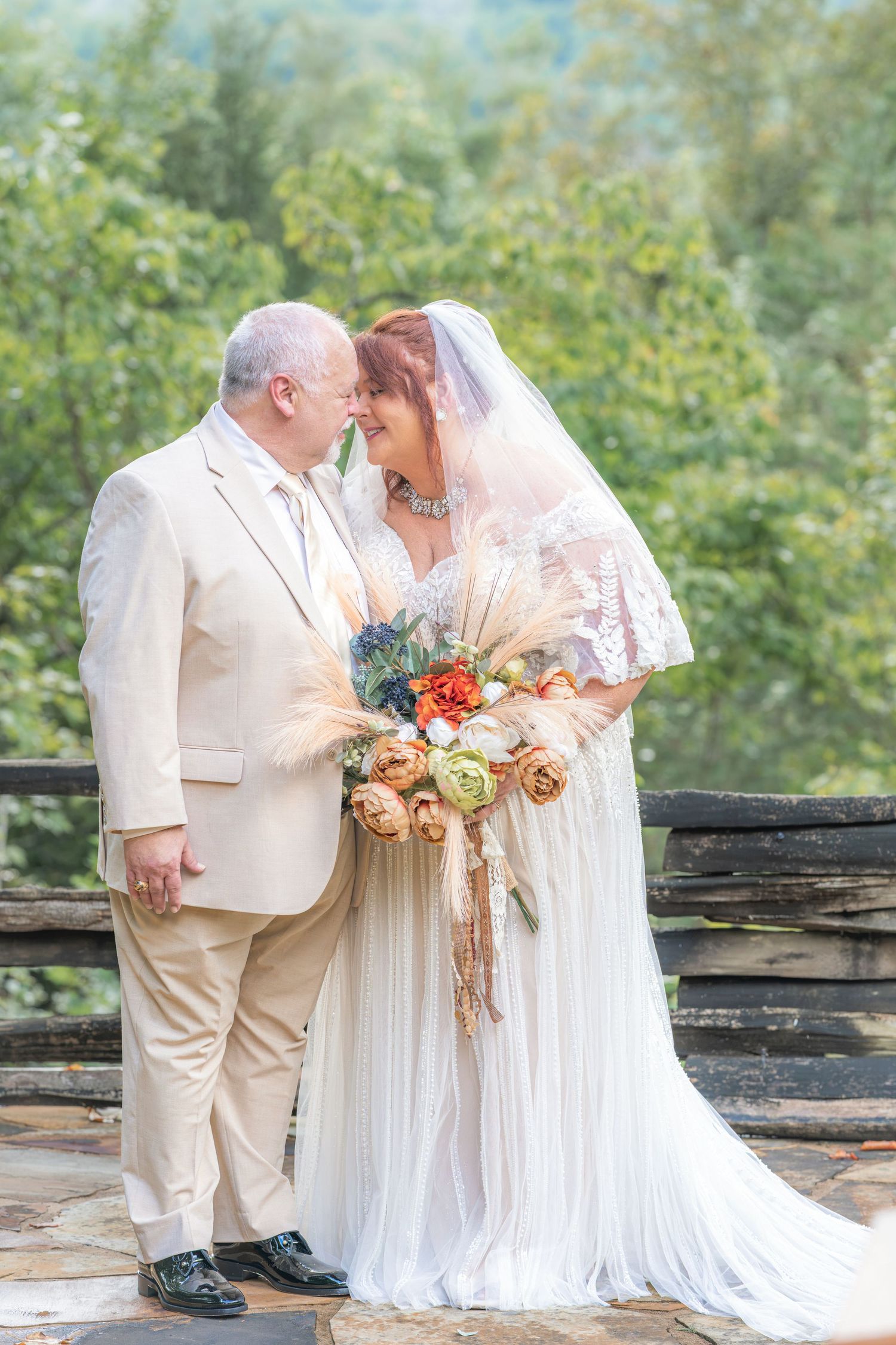 Mature bride and groom in cream wedding attire share a romantic moment outdoors, bride holding a colorful bouquet.