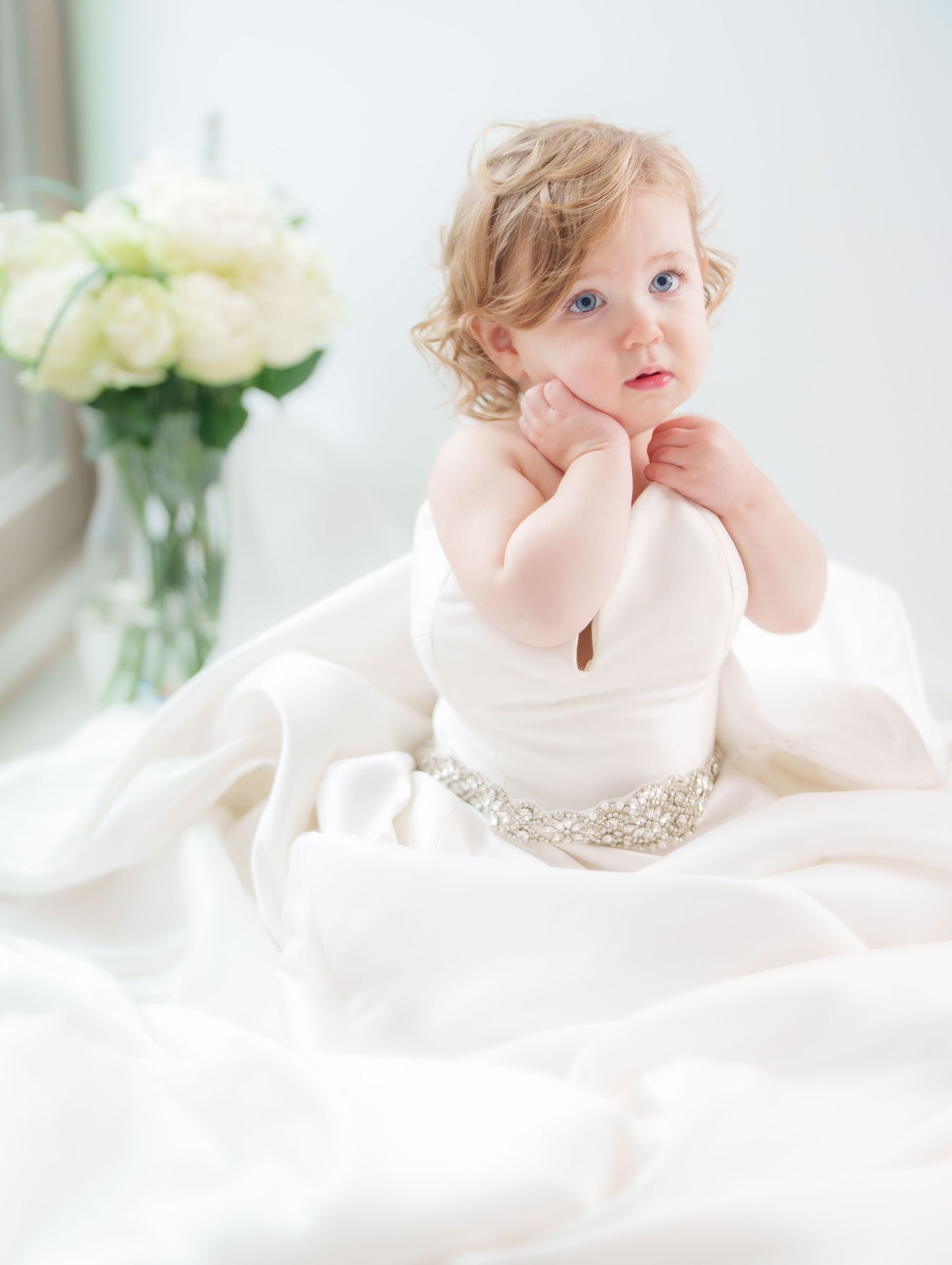Adorable blue-eyed toddler sitting in a white wedding gown with a rhinestone belt.