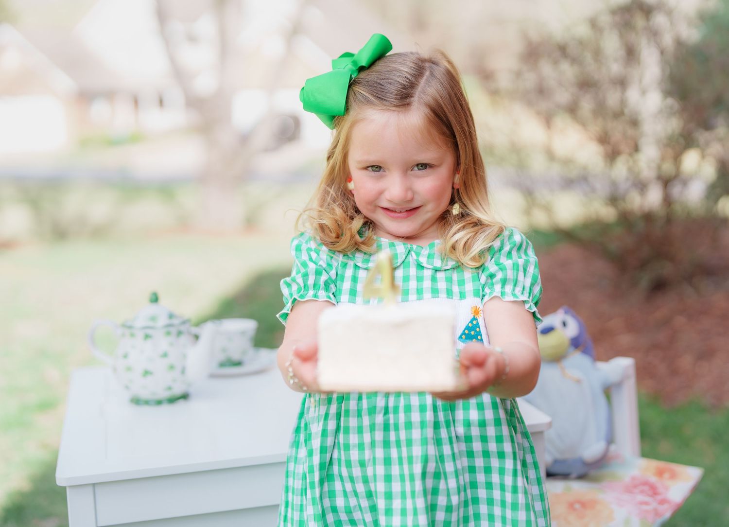 A smiling young girl in a green gingham dress with a green bow holds out a white gift box outdoors.