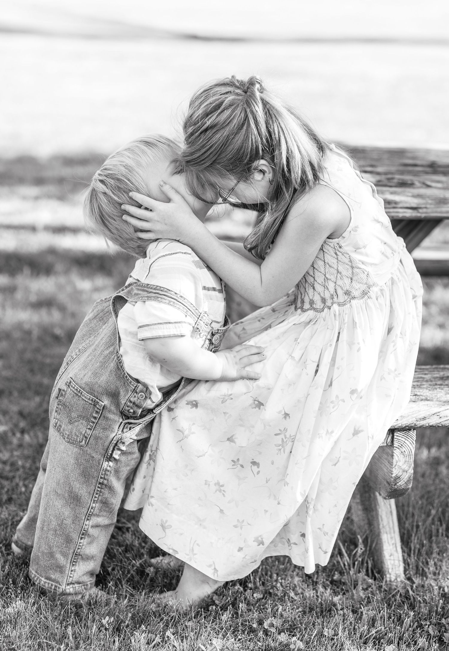 Black and white photo of a young girl hugging a toddler boy on a bench outdoors.