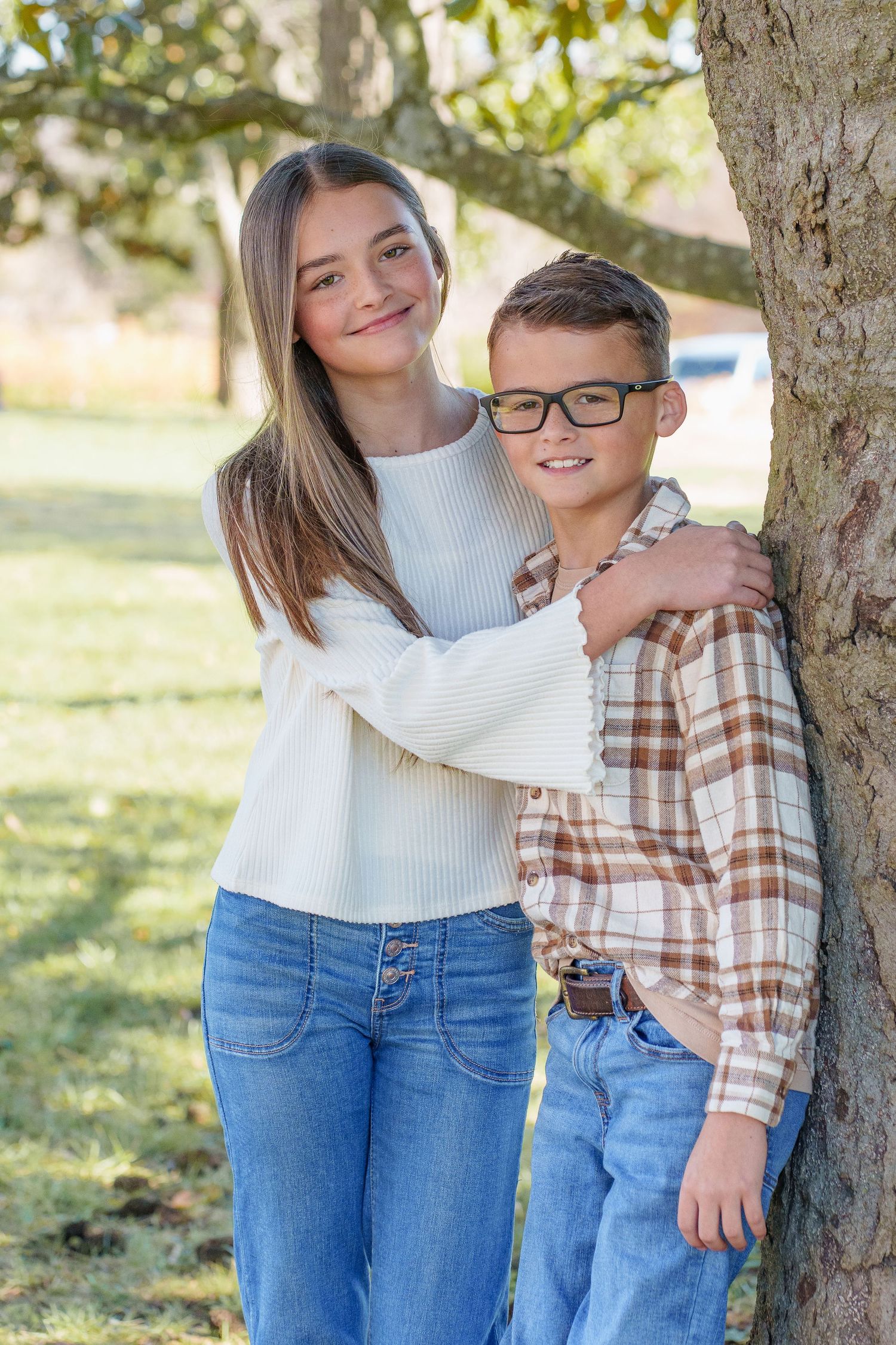 Teenage girl hugging younger brother with glasses leaning against tree in park.