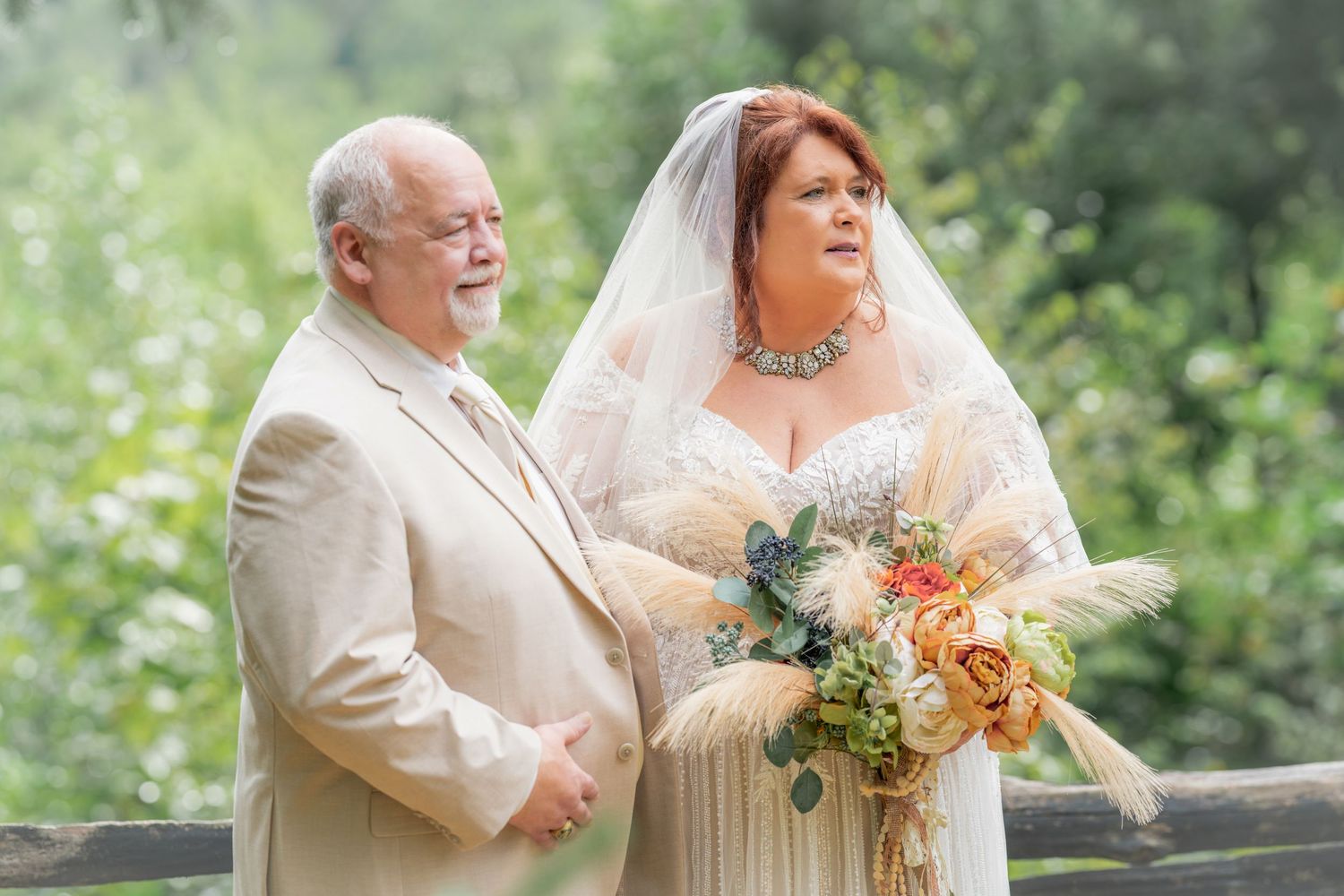 Bride in lace gown and veil holds bouquet beside man in tan suit at outdoor wedding ceremony in Gatlinburg, TN.