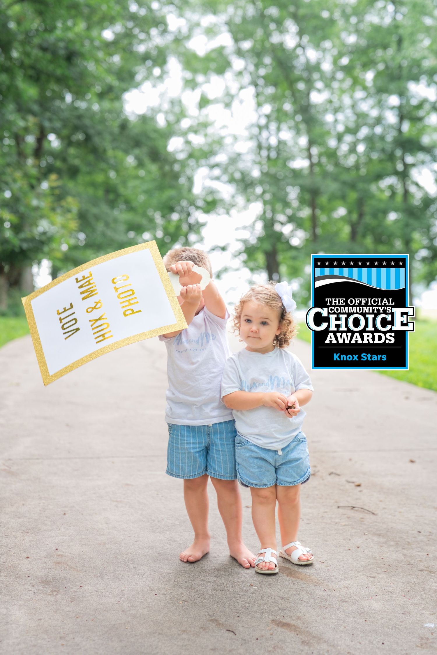 Two young children in white shirts hold a voting sign outdoors, promoting Hux & Mae Photo for Community Choice Awards.