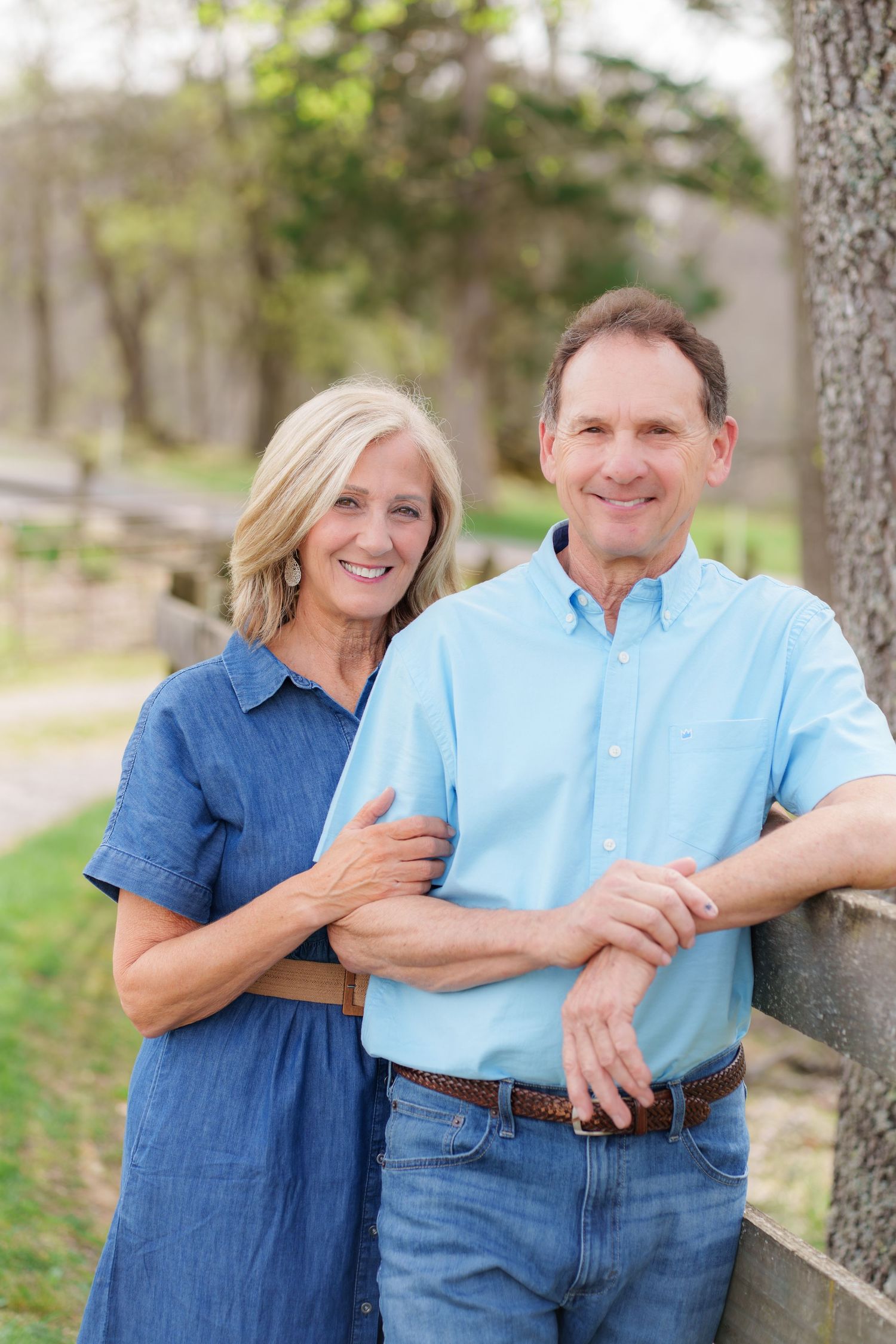 Smiling middle-aged couple posing outdoors near a wooden fence and tree in a lush green park setting.